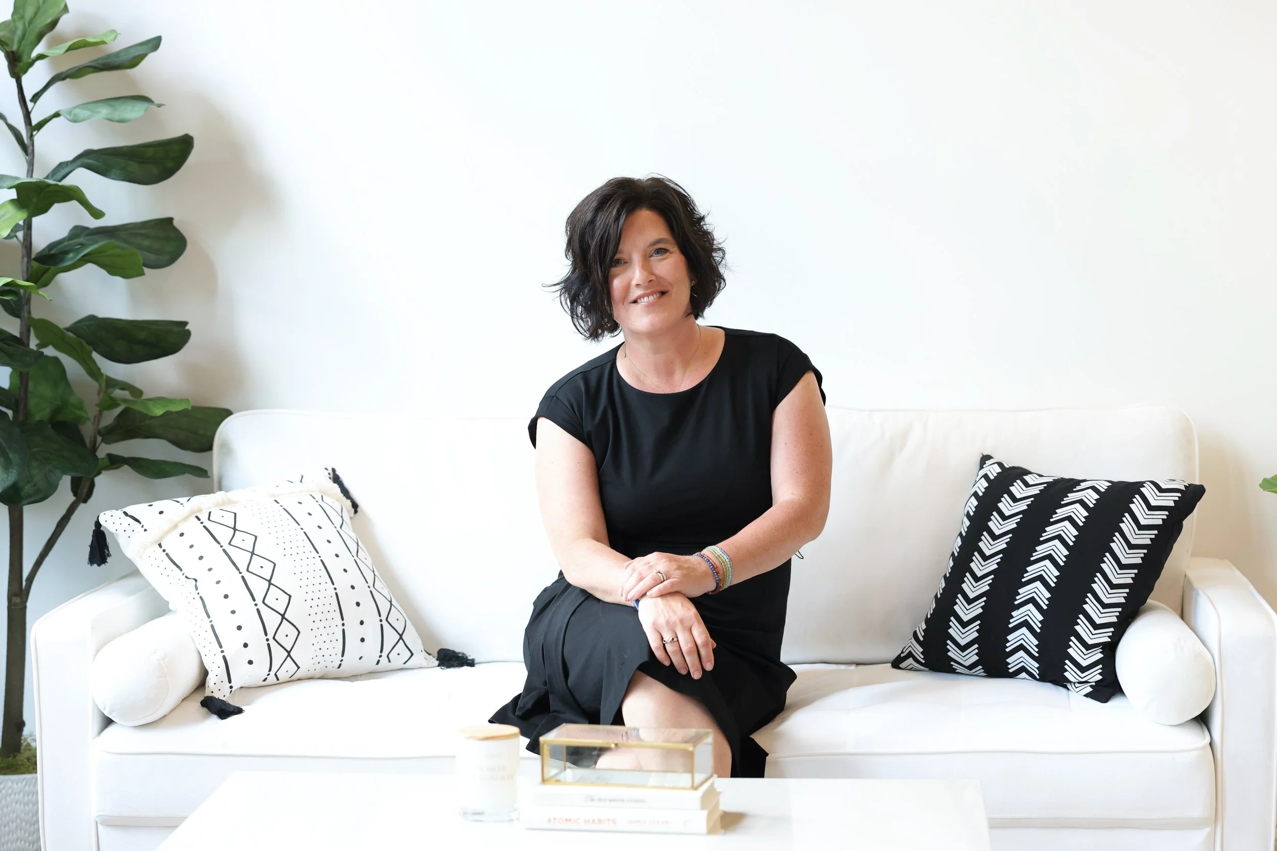 Angela Gowans with short dark wavy hair sitting on a white sofa with black and white patterned pillows, in a bright minimalist living room with green plants, white walls, and a coffee table with books and a candle.
