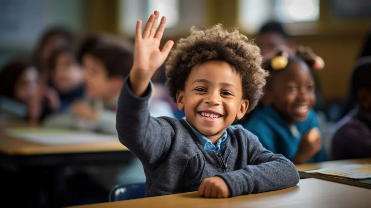 Young boy with curly hair smiling and raising his hand in a classroom full of children.