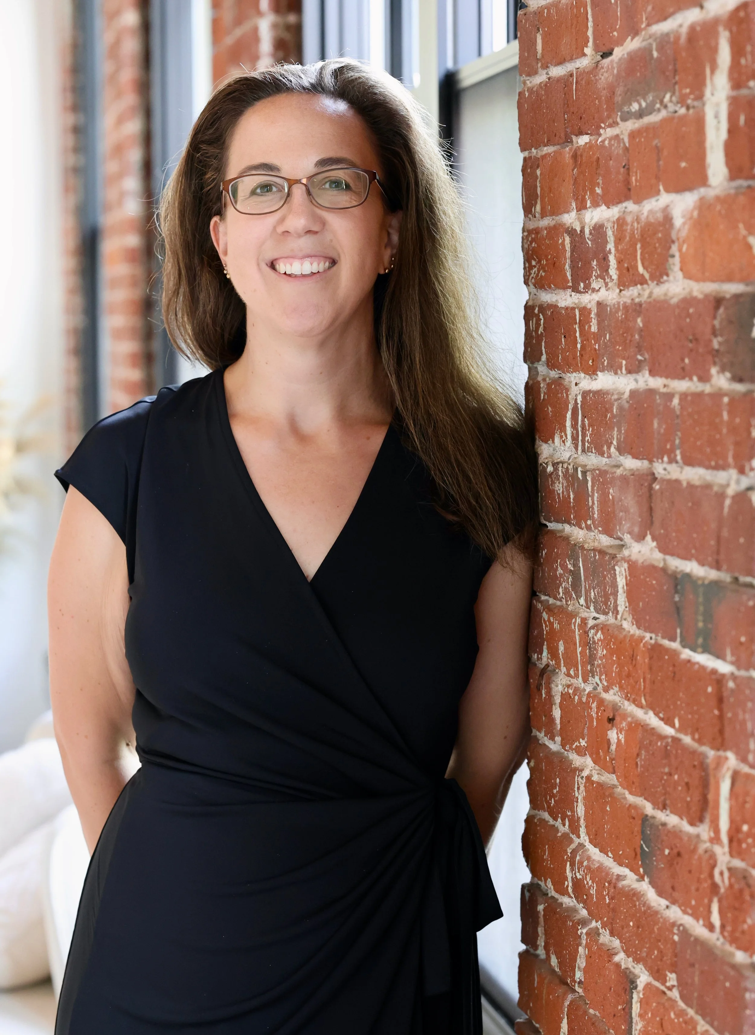 Emily Dean with long brown hair, glasses, and wearing a black dress, standing next to a brick wall inside a building with large windows.