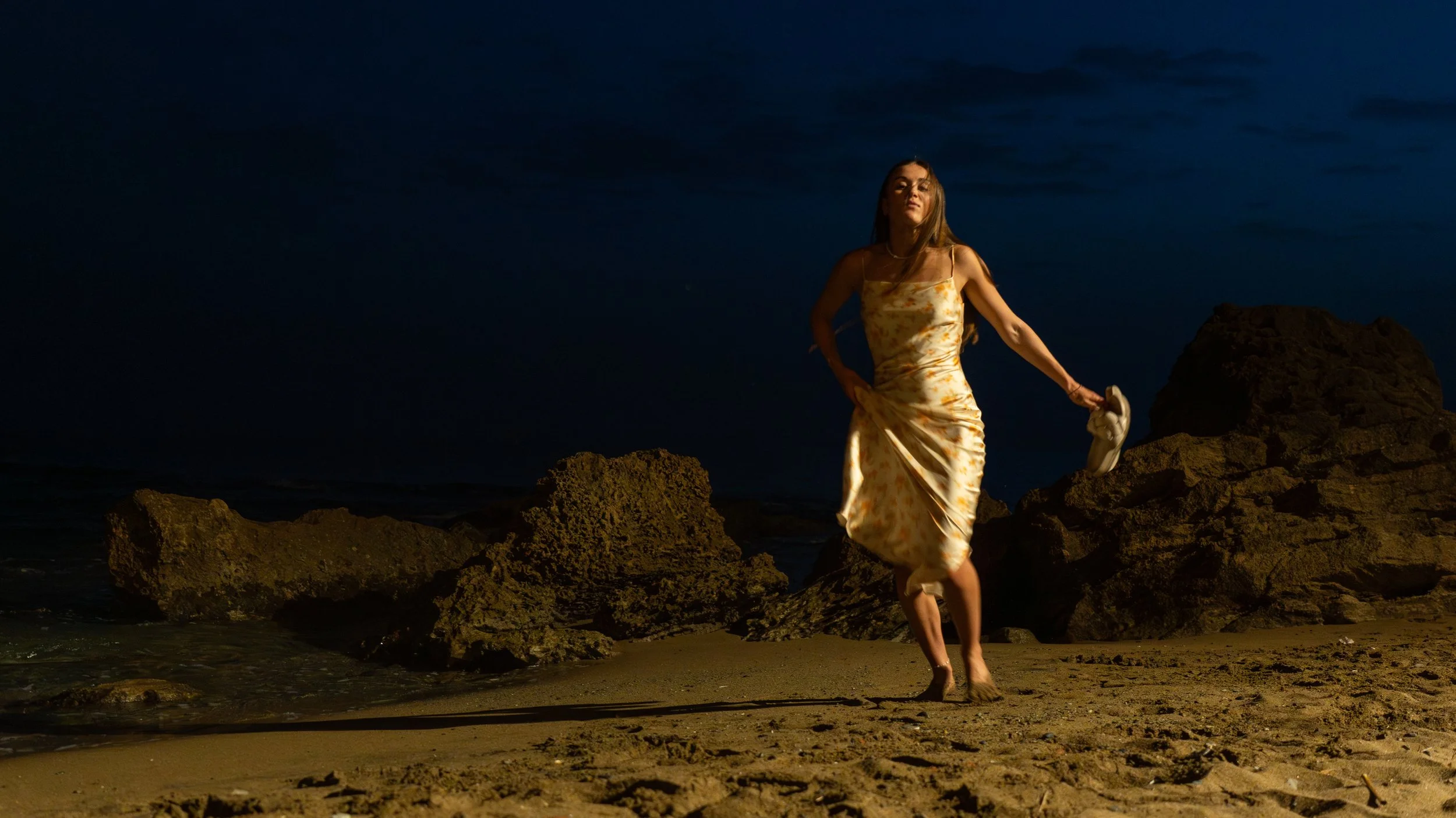 A woman in a floral dress walking barefoot on rocky beach at night, holding shoes, with dark sky and ocean in the background.