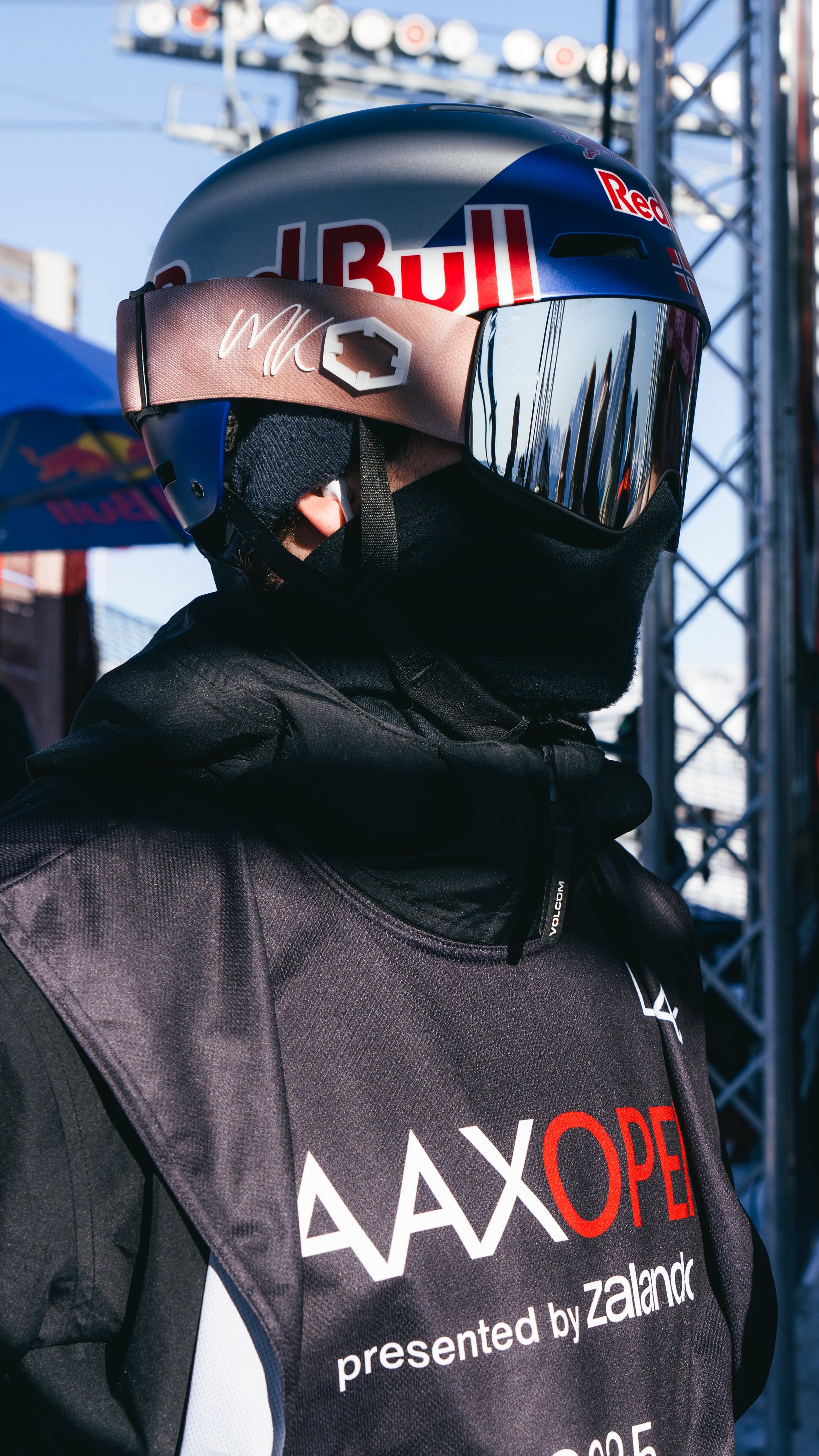 Close-up of a person wearing a helmet with Red Bull branding and mirrored visor, dressed in black gear with a vest bearing logos, at an outdoor event with metal structures in the background.