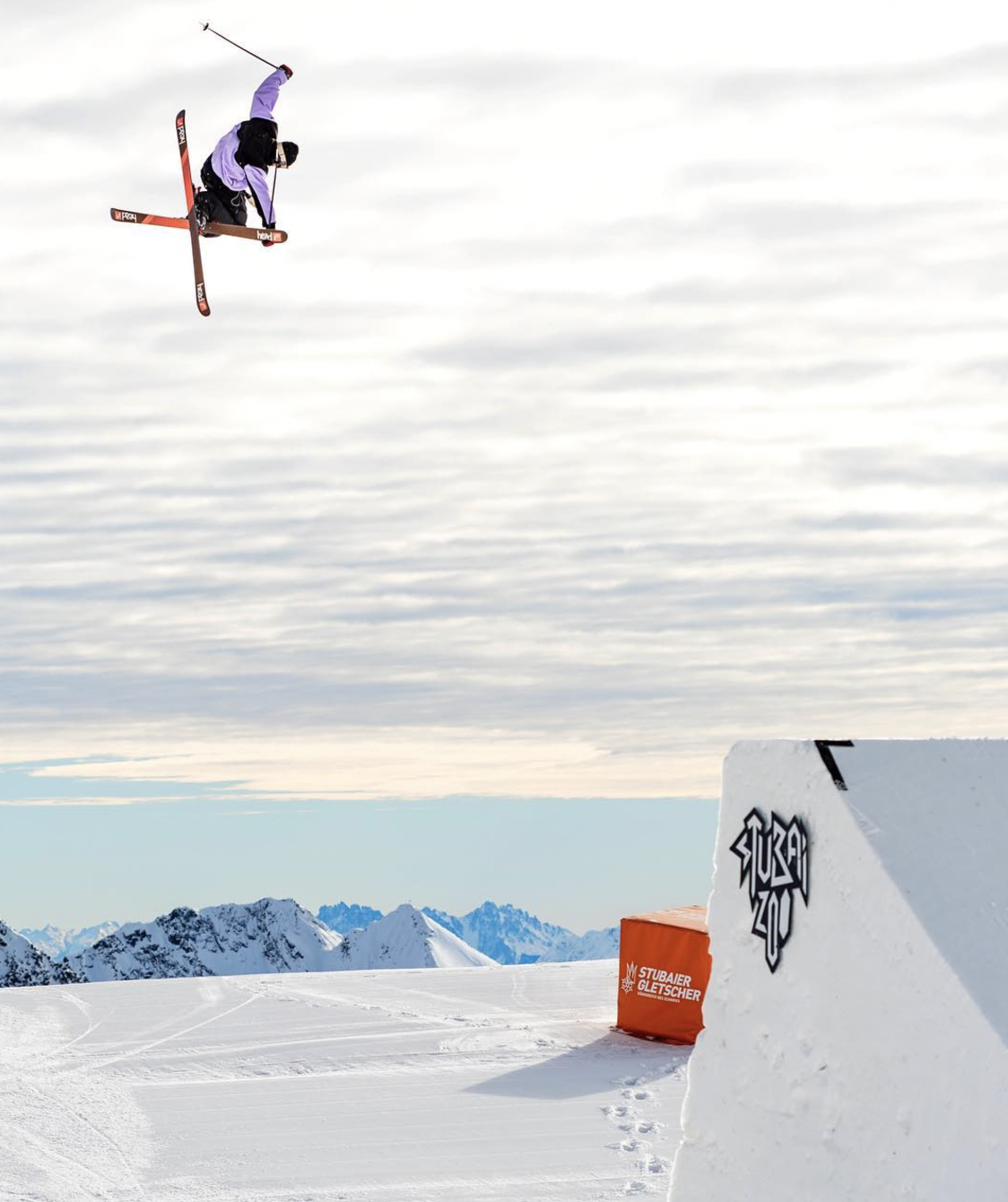 A skier performing a flip in mid-air on a snowy ski jump ramp with mountains in the background