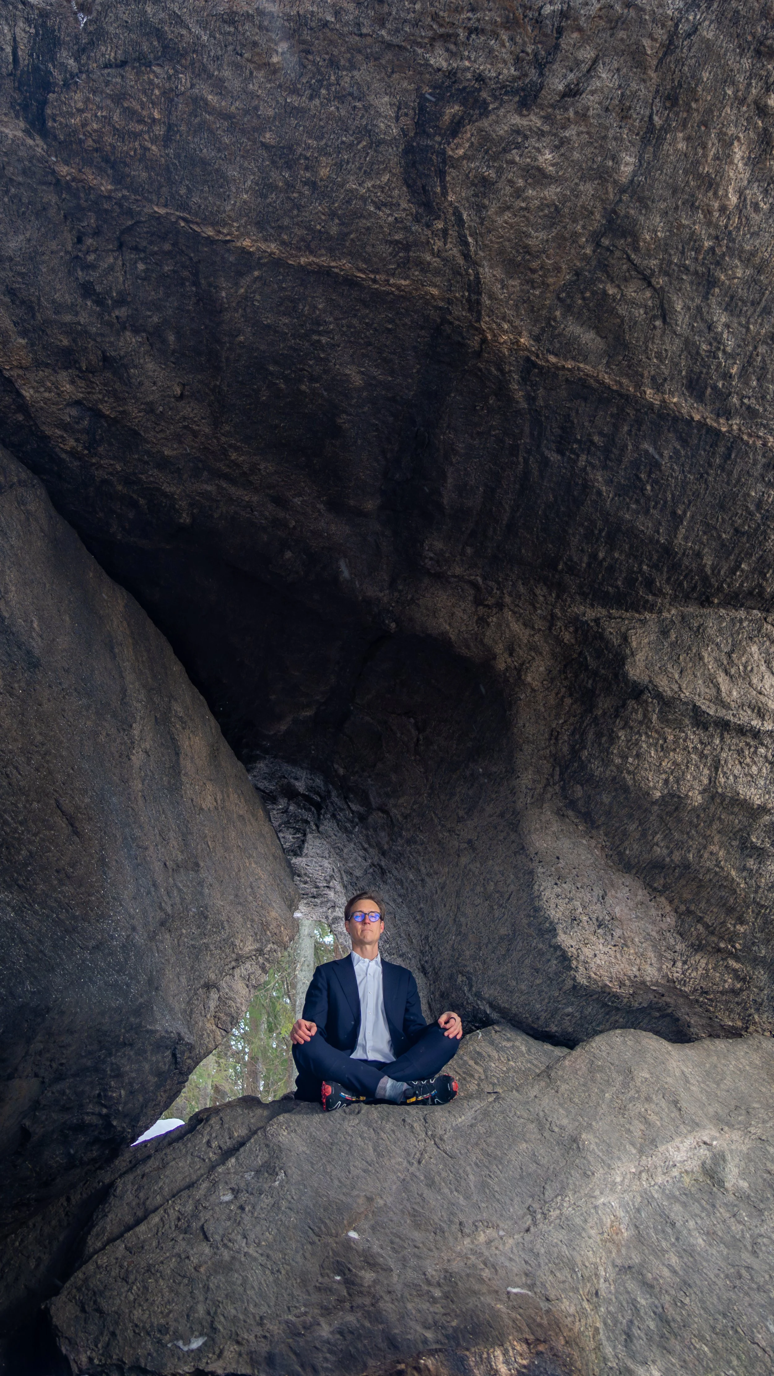 A man in a suit and glasses meditating in a seated cross-legged position inside a large rock formation with an opening above, surrounded by trees.