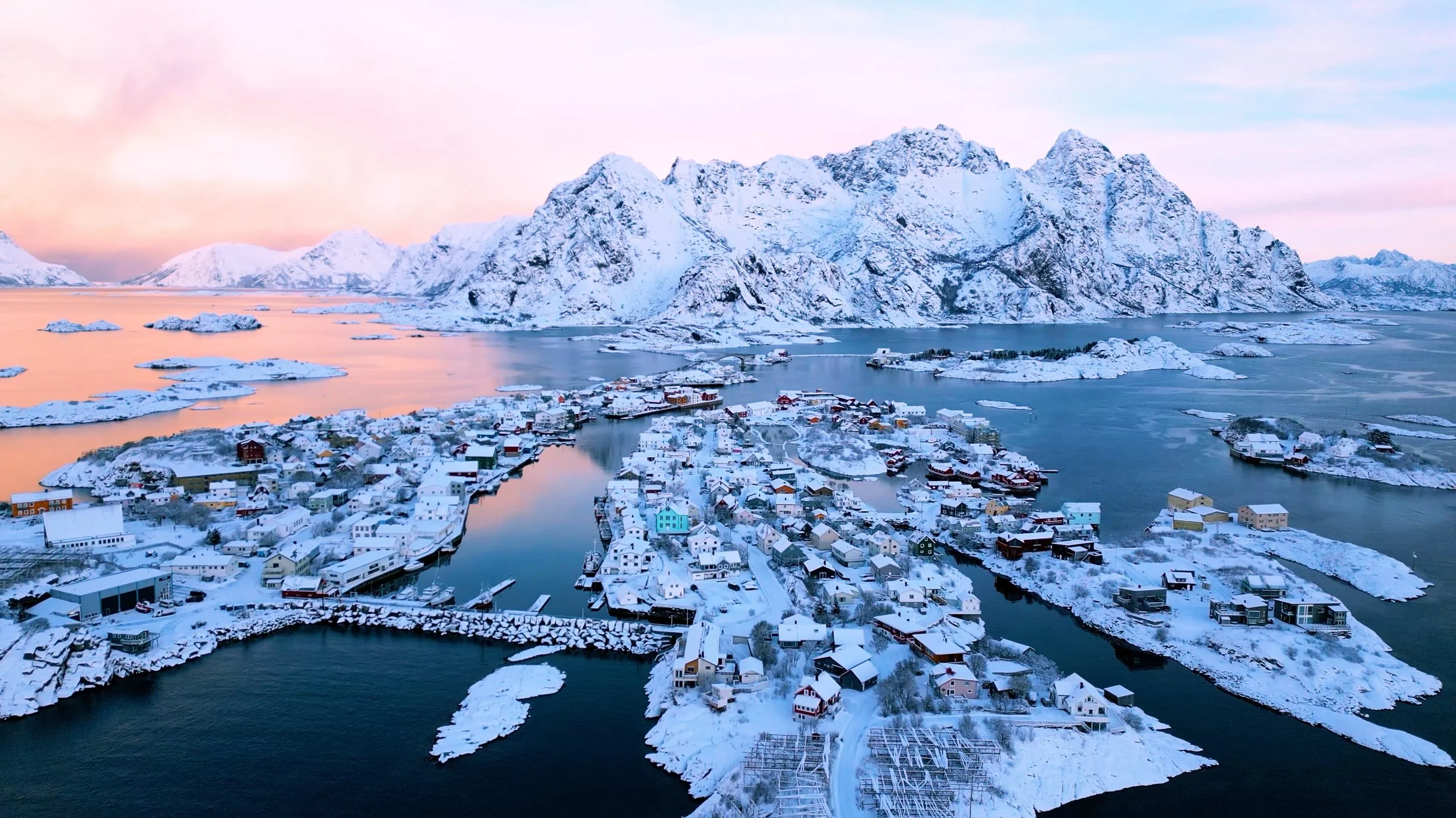 Snow-covered village with colorful houses along a fjord, surrounded by icy mountains at sunset.