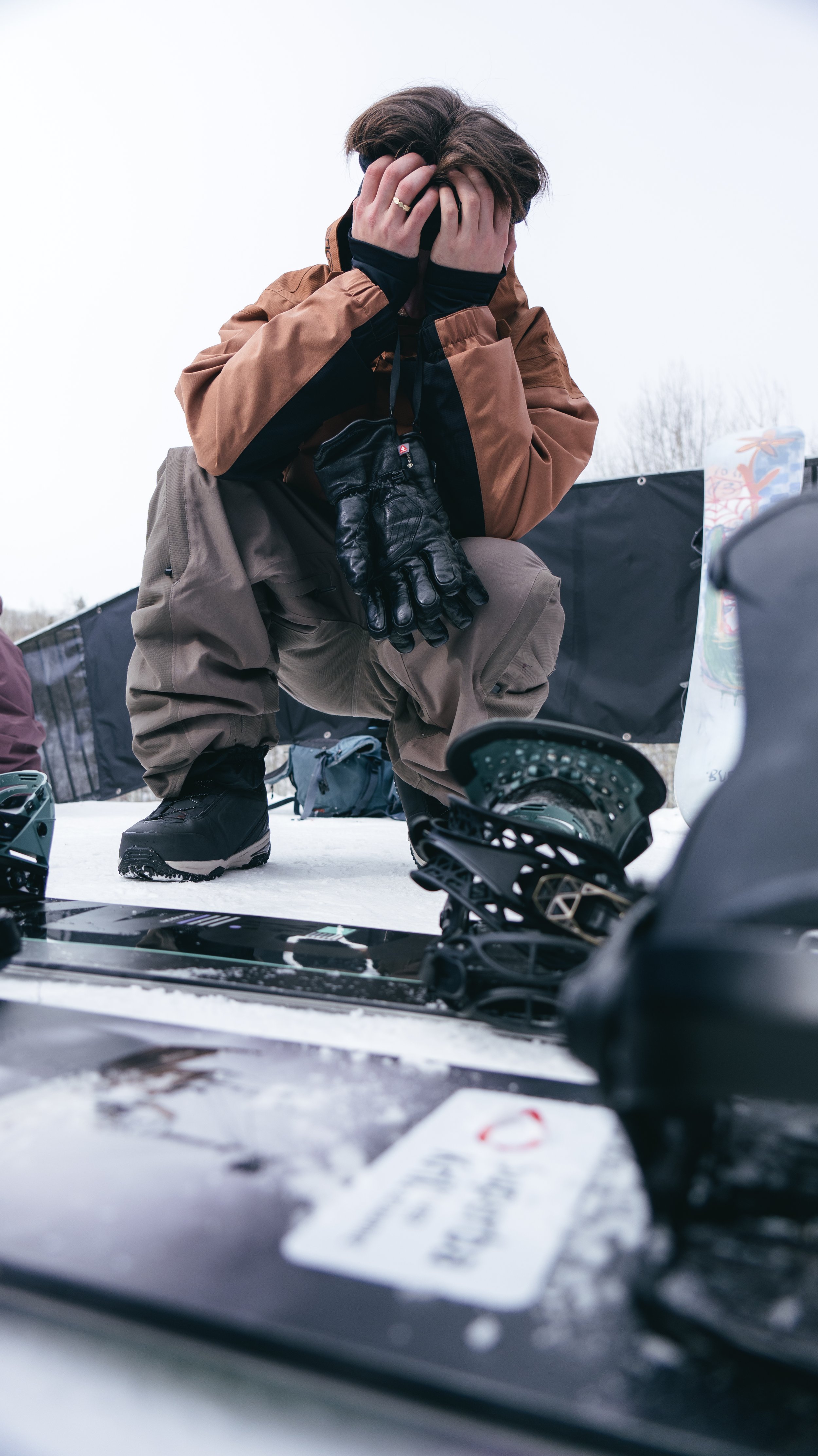 Person crouching outdoors in snowy weather with hands covering their face, surrounded by snowboarding gear, including skis and a snowboard.