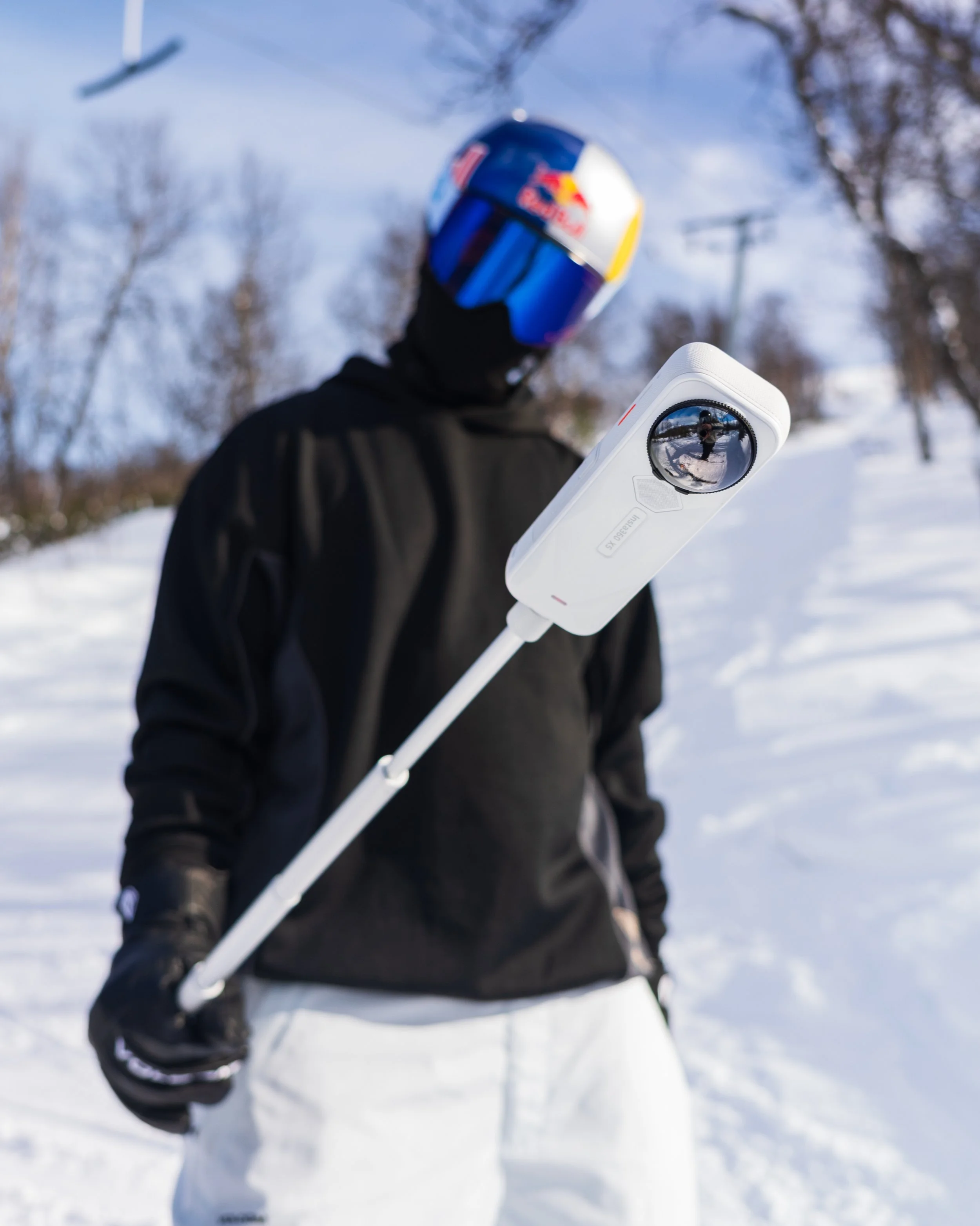 A person wearing a blue helmet with a Red Bull logo, black winter gear, and holding a white ski pole with a reflective mirror, standing on snow with a winter landscape background.