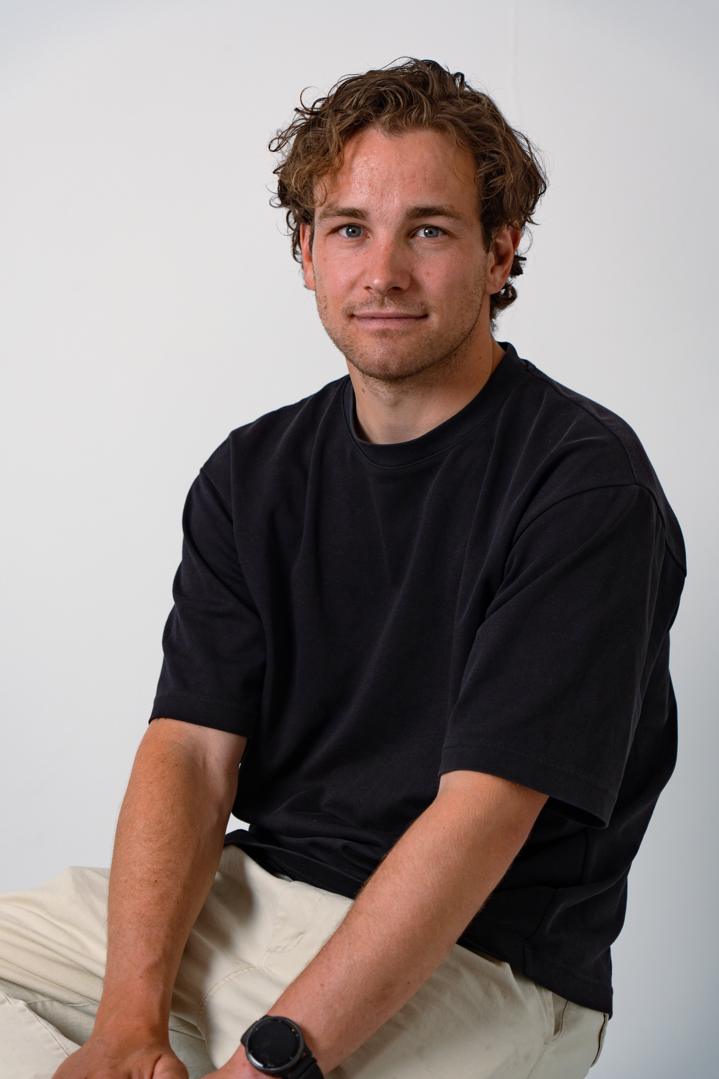 A young man with wavy brown hair and blue eyes wearing a black t-shirt and a black watch, sitting against a plain white background.