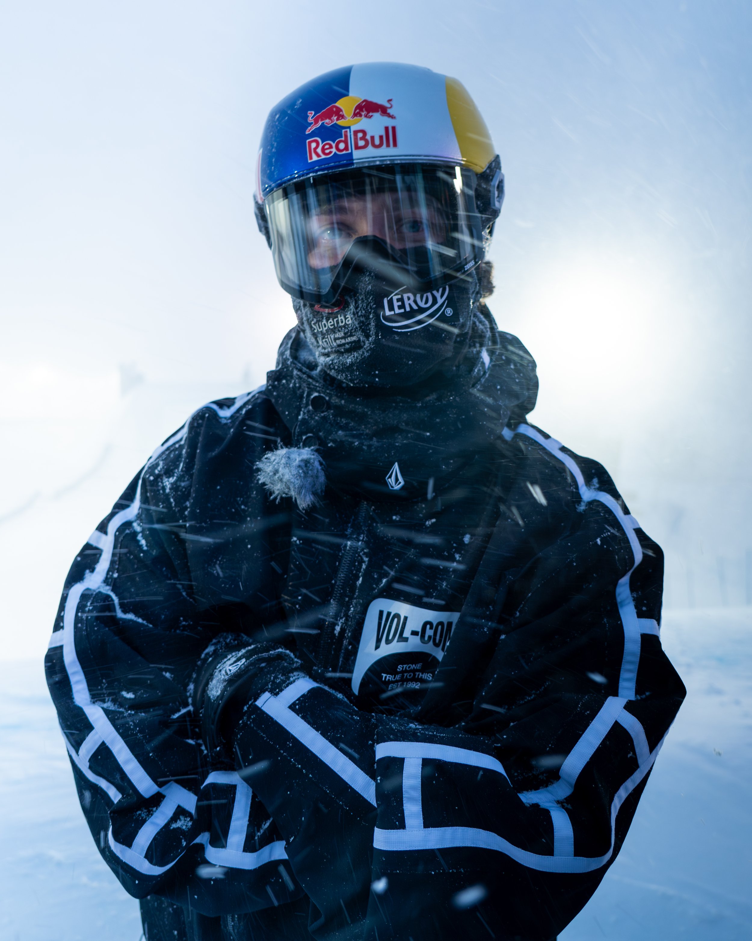 Person wearing a Red Bull racing helmet and black jacket with white and blue accents, standing in snowy environment with snow falling.