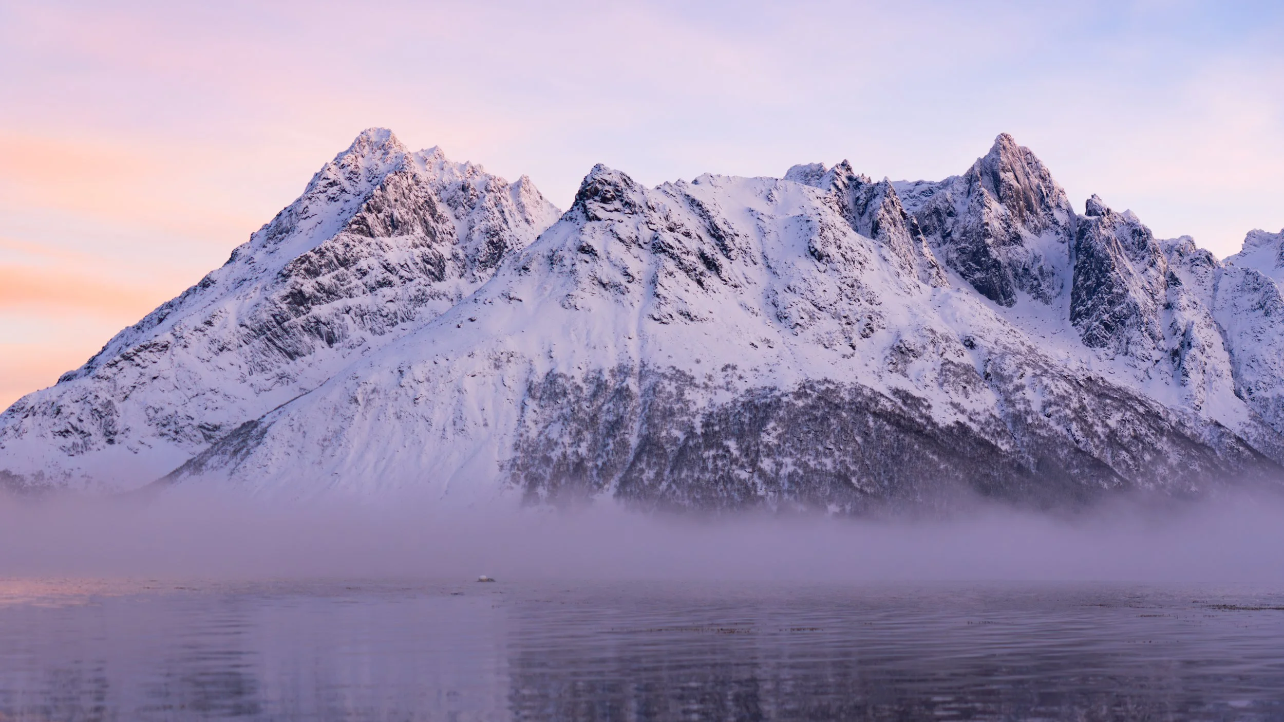Snow-covered mountain range beside a body of water during dusk or dawn with pastel-colored sky.
