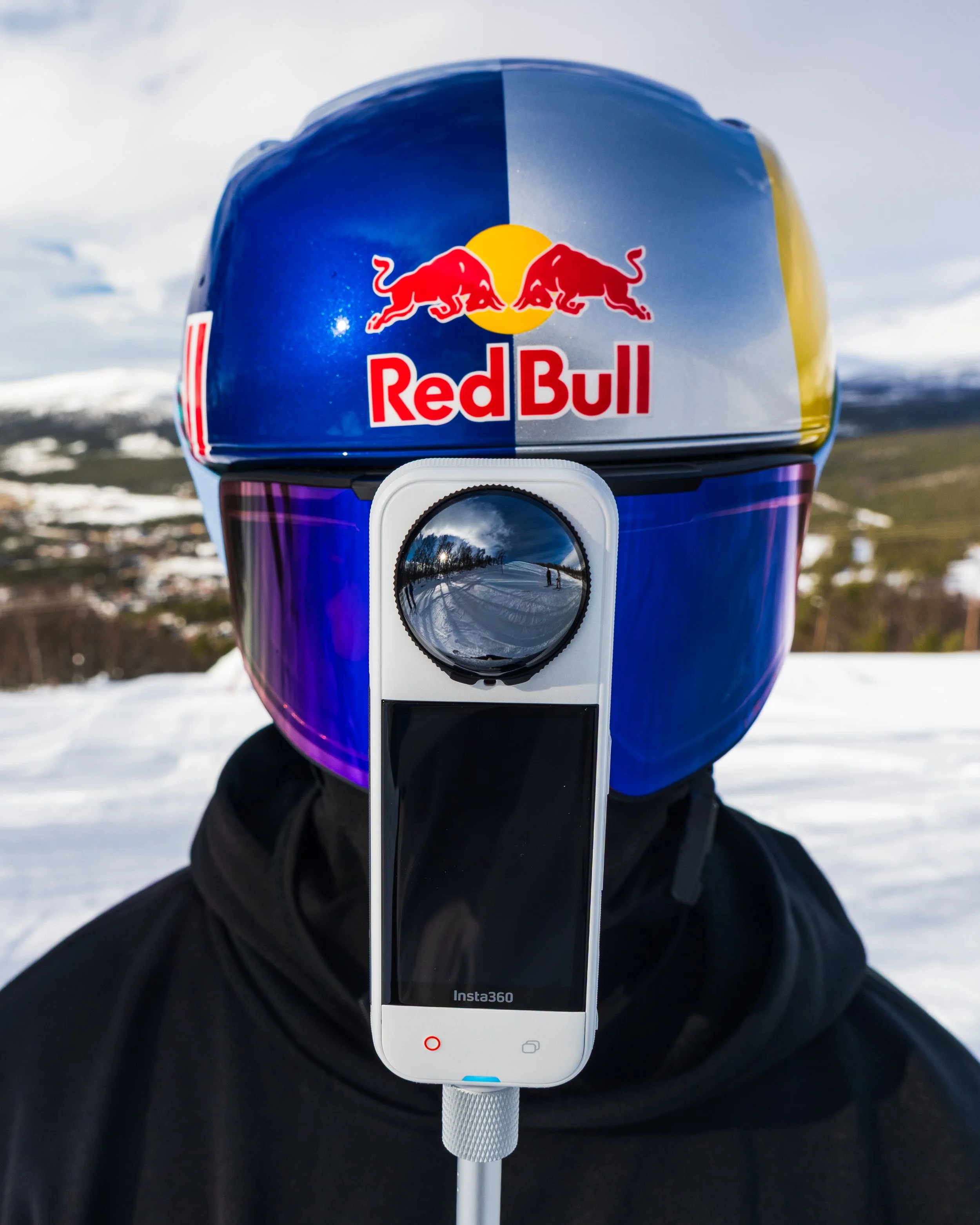 Close-up of a motorcycle helmet with a Red Bull logo, mounted on a selfie stick with a smartphone showing the brand Insta360, against a snowy landscape background.