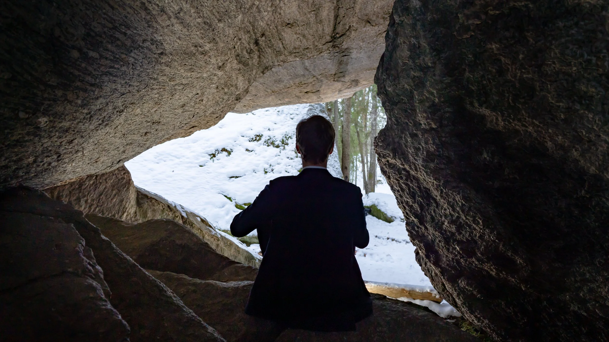 Person in a suit sitting inside a rocky cave opening, looking out at snow-covered ground and trees.