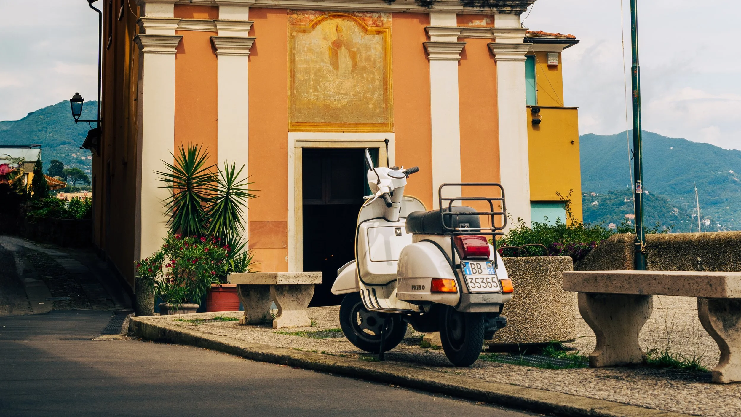 A small white scooter parked in front of a peach-colored building with white architectural details and a faded religious fresco above the doorway. There are plants in pots and a stone bench nearby, with mountains in the background.