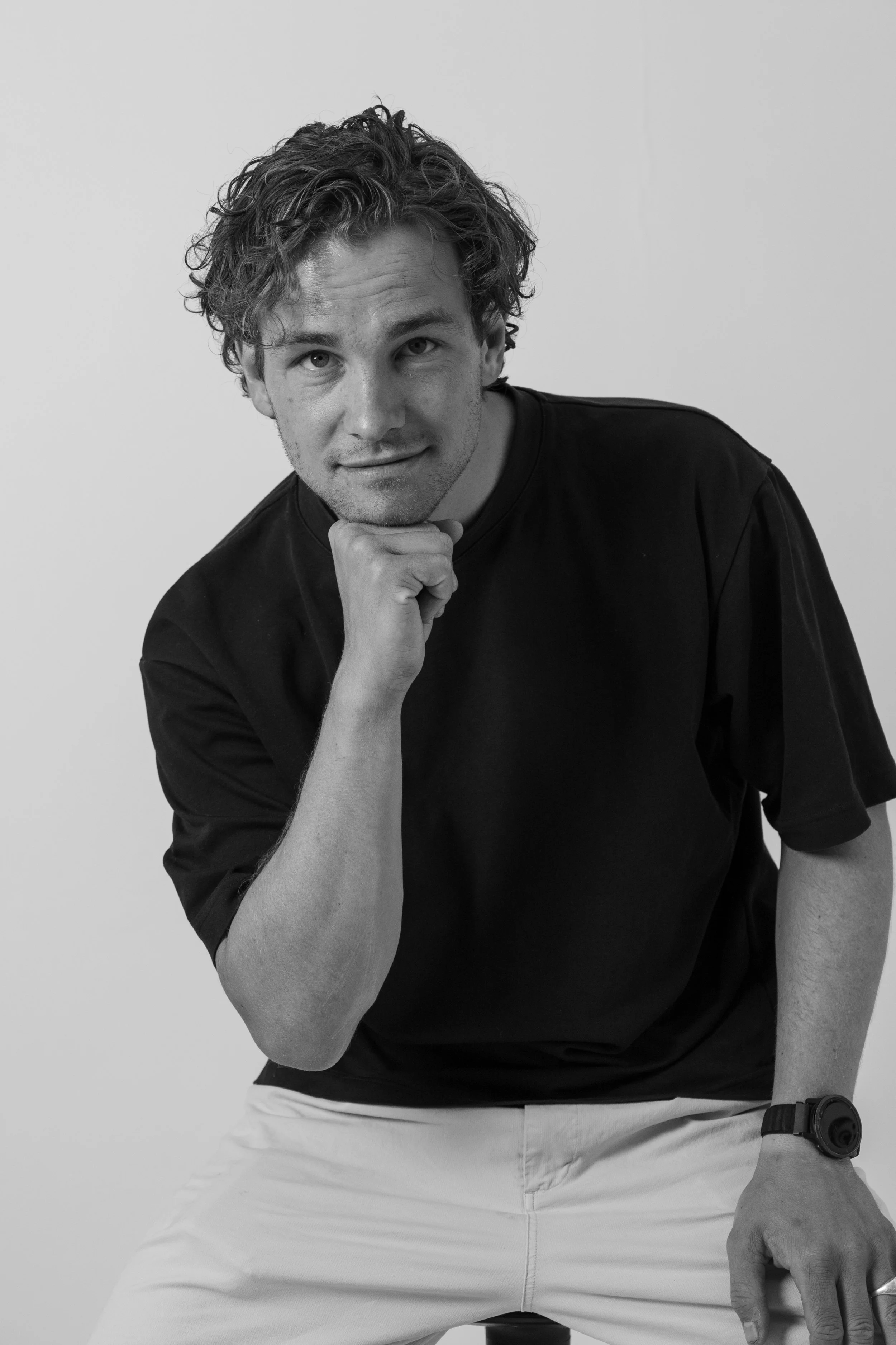 Black and white photo of a man with curly hair, resting his chin on his left hand, sitting on a chair, wearing a black shirt, white pants, and a watch on his right wrist, against a plain background.