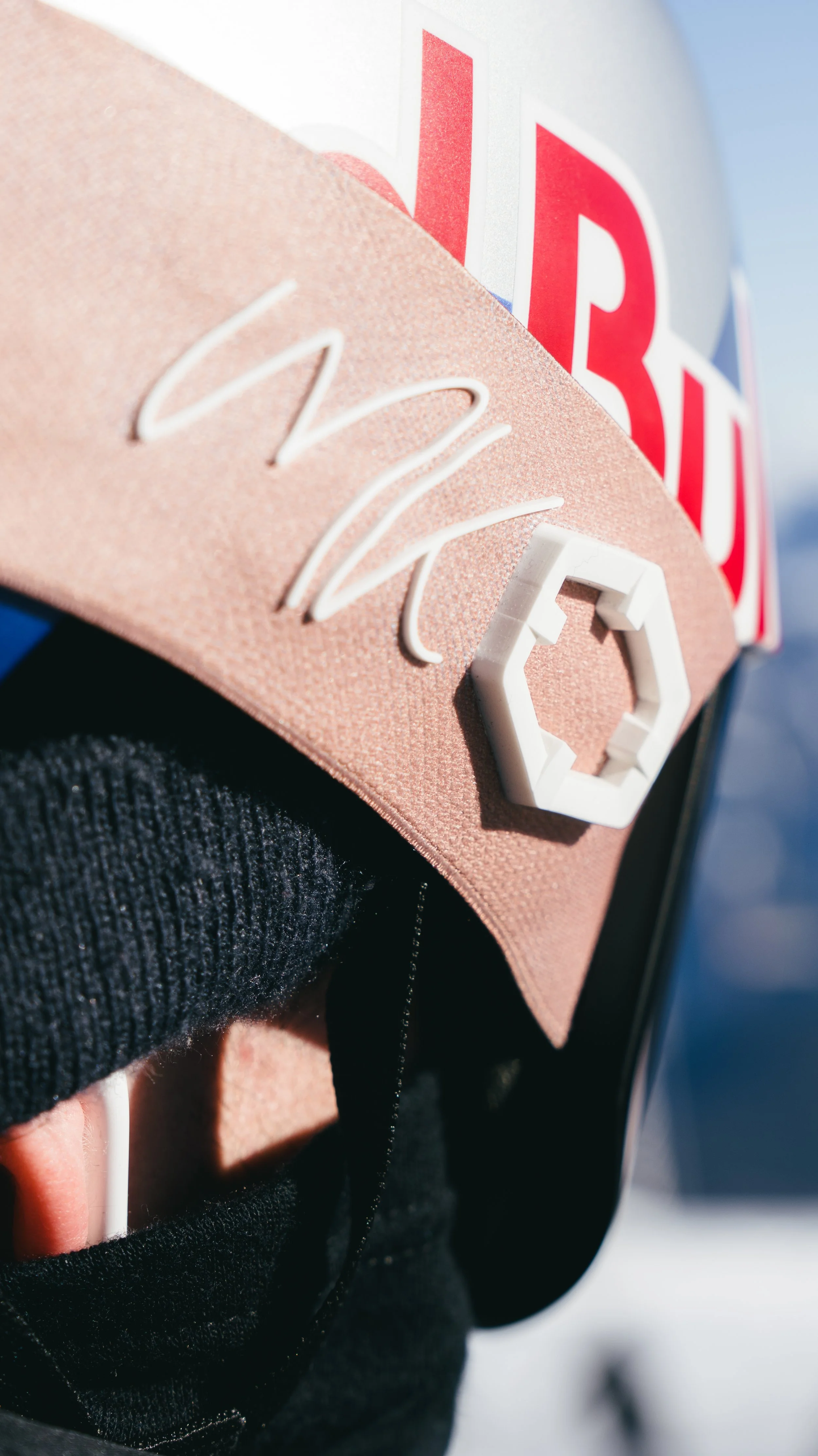 Close-up of a person in a black jacket with a knit cuff, wearing a pinkish cap with a white 3D geometric and neon sign logo, and a partially visible helmet with red, white, and blue text.
