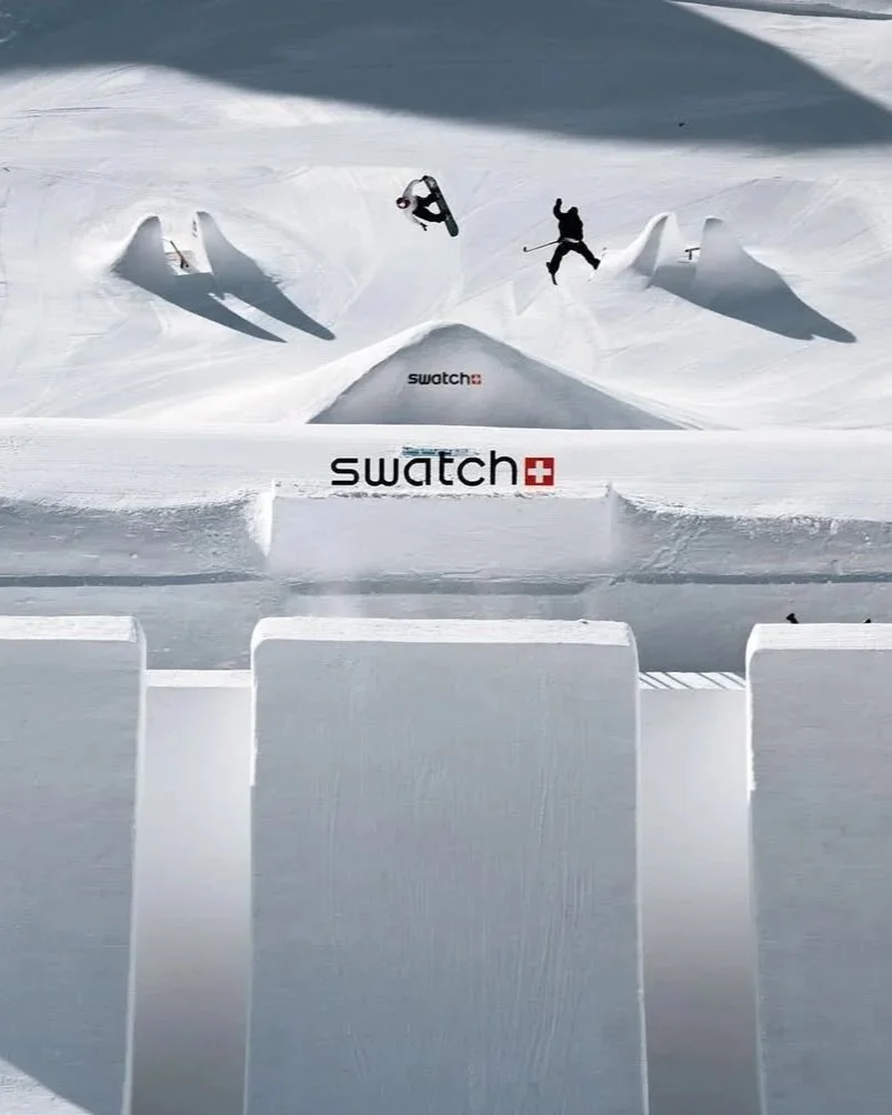A skier performing tricks in a snow park with large jumps and rails, with Swatch branding visible on the snow.