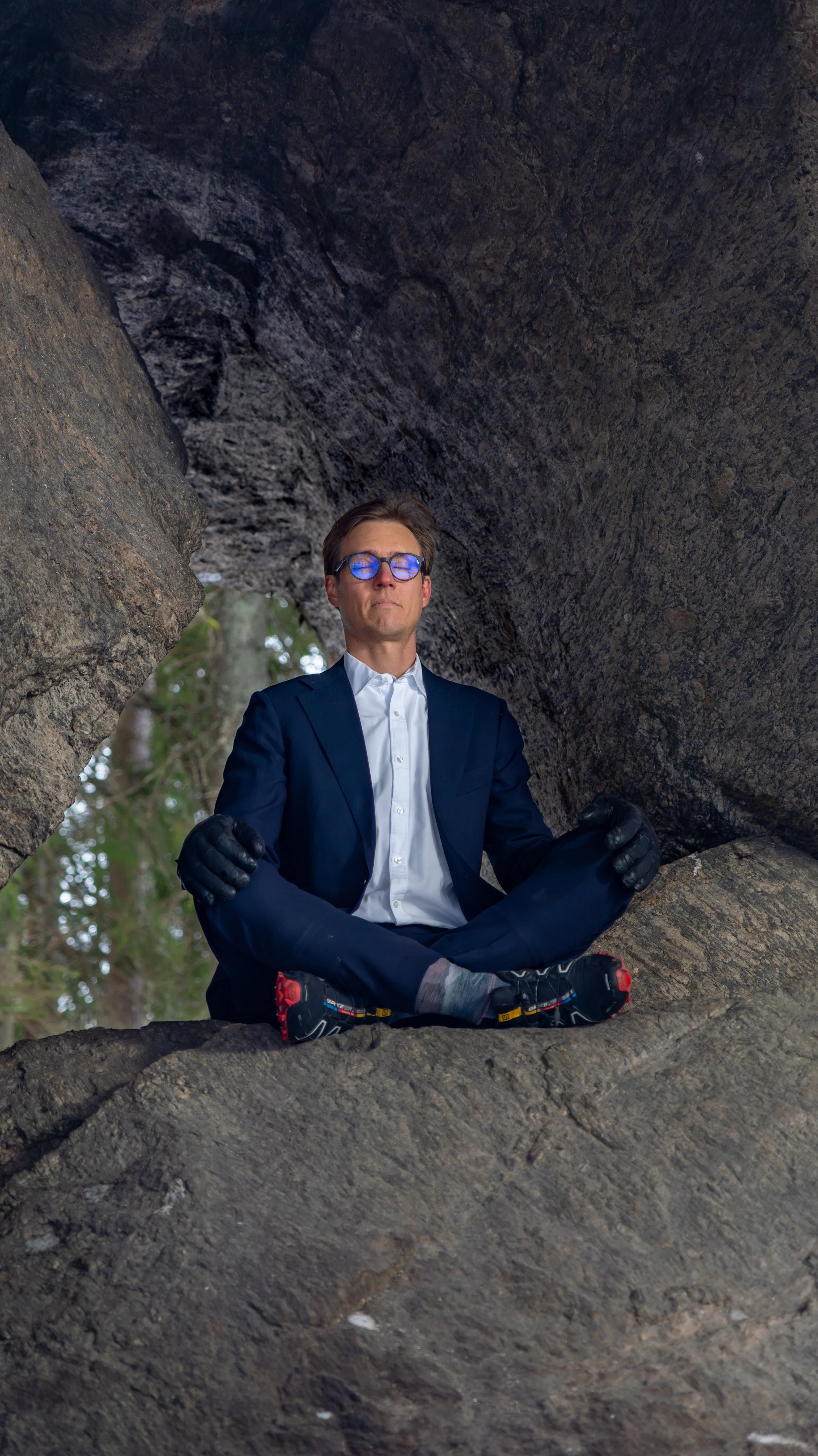 A man in sunglasses, a formal suit, black gloves, and sneakers is sitting cross-legged inside a rock formation, with trees visible in the background.