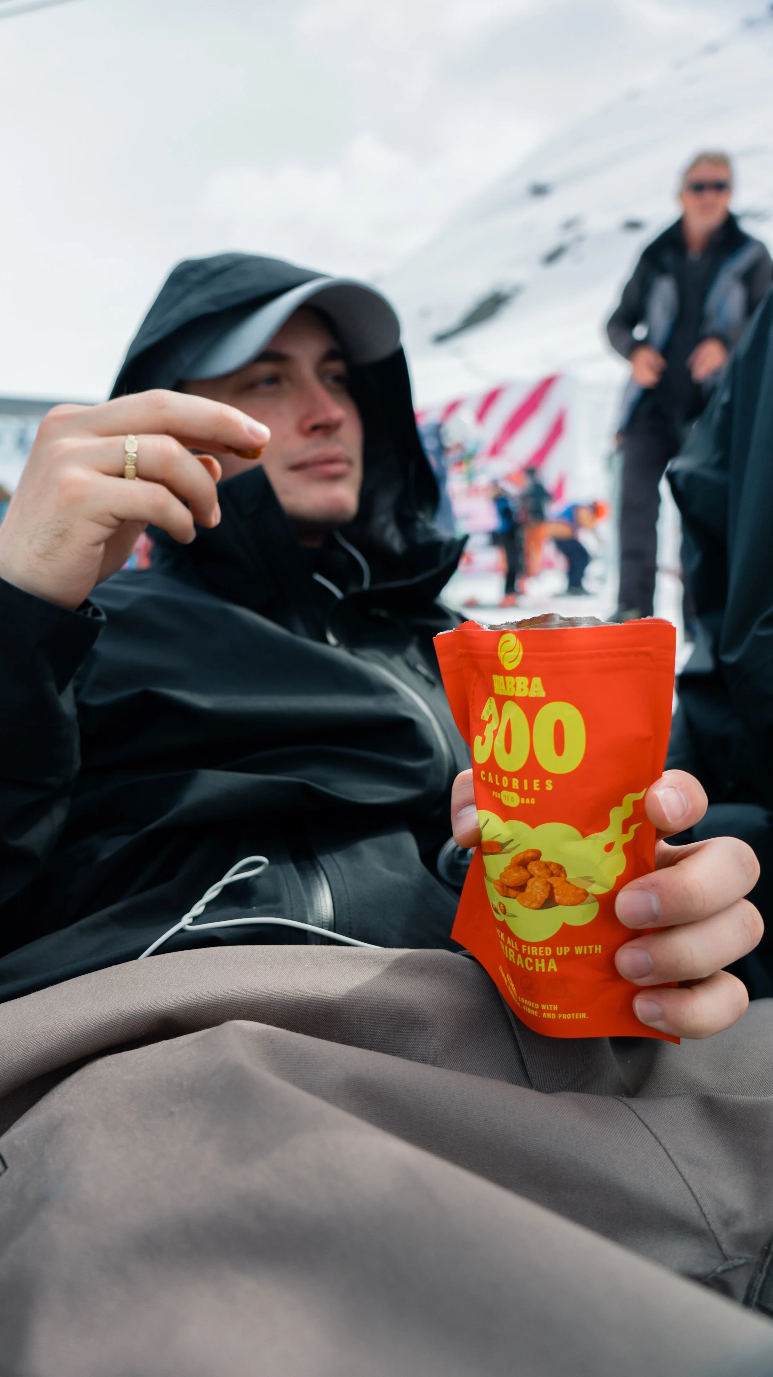 A man wearing a hoodie and jacket sitting outdoors, holding a bag of spicy flavored snack with the label '300 calories.' He appears to be in a snowy mountain area, with snow-covered peaks in the background and a person standing in the distance.