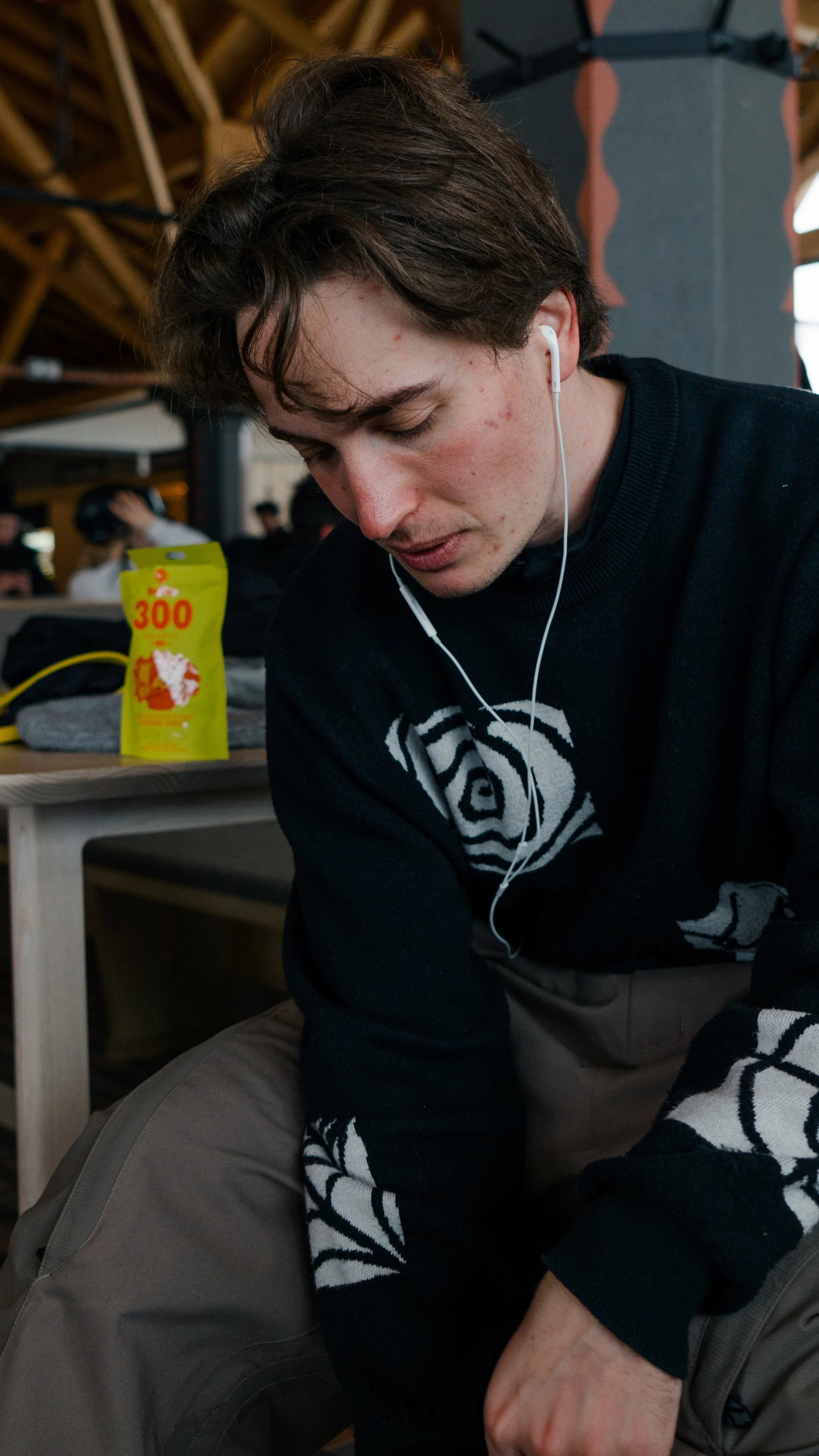 A young man with brown hair and light skin, wearing a black sweater with a white patterned design, is sitting and listening to earphones in a busy indoor space. There is a yellow snack bag on a table behind him.