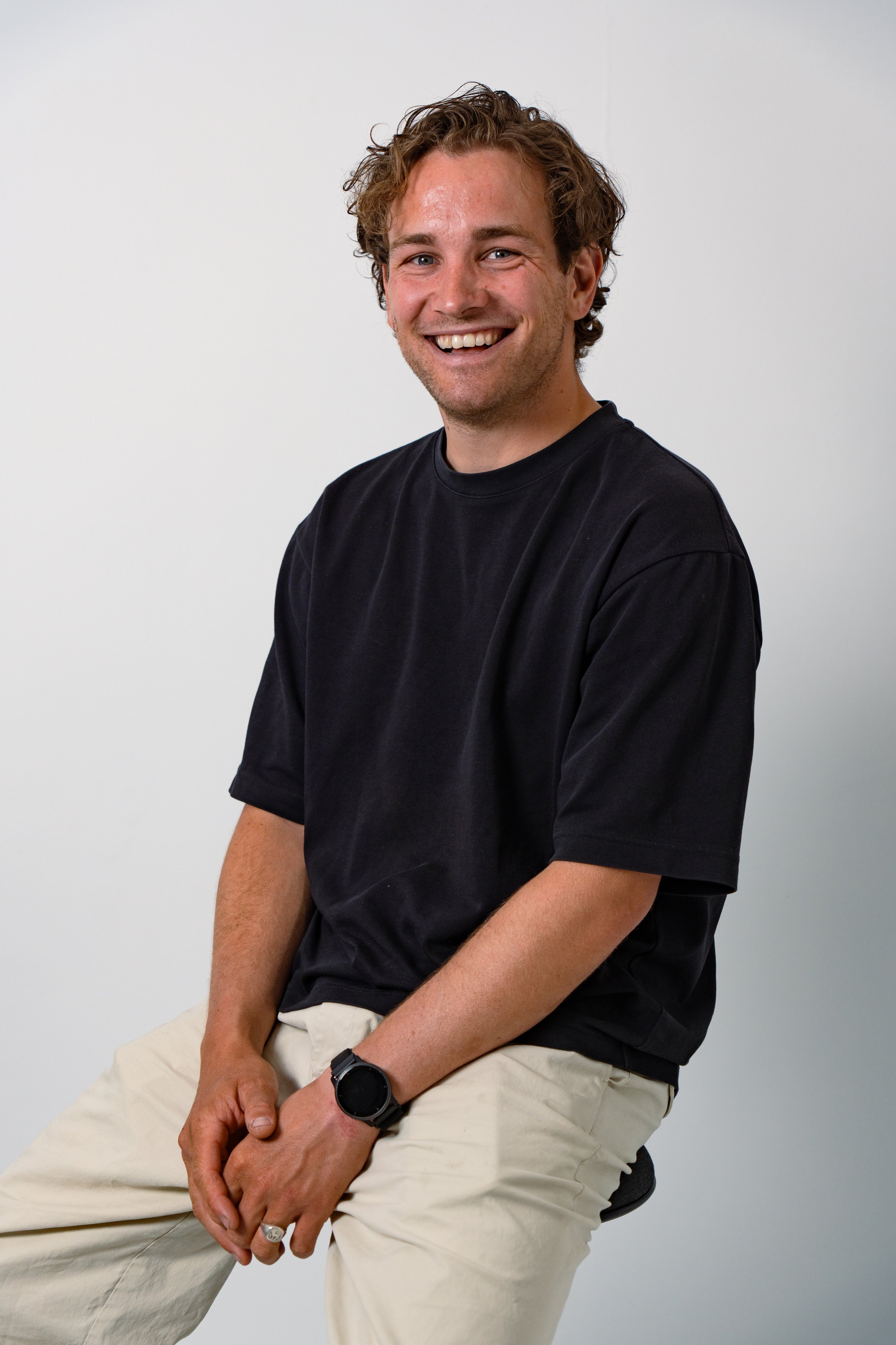 A young man with curly brown hair, smiling, wearing a black T-shirt, beige pants, and a black wristwatch, sitting against a plain white background.