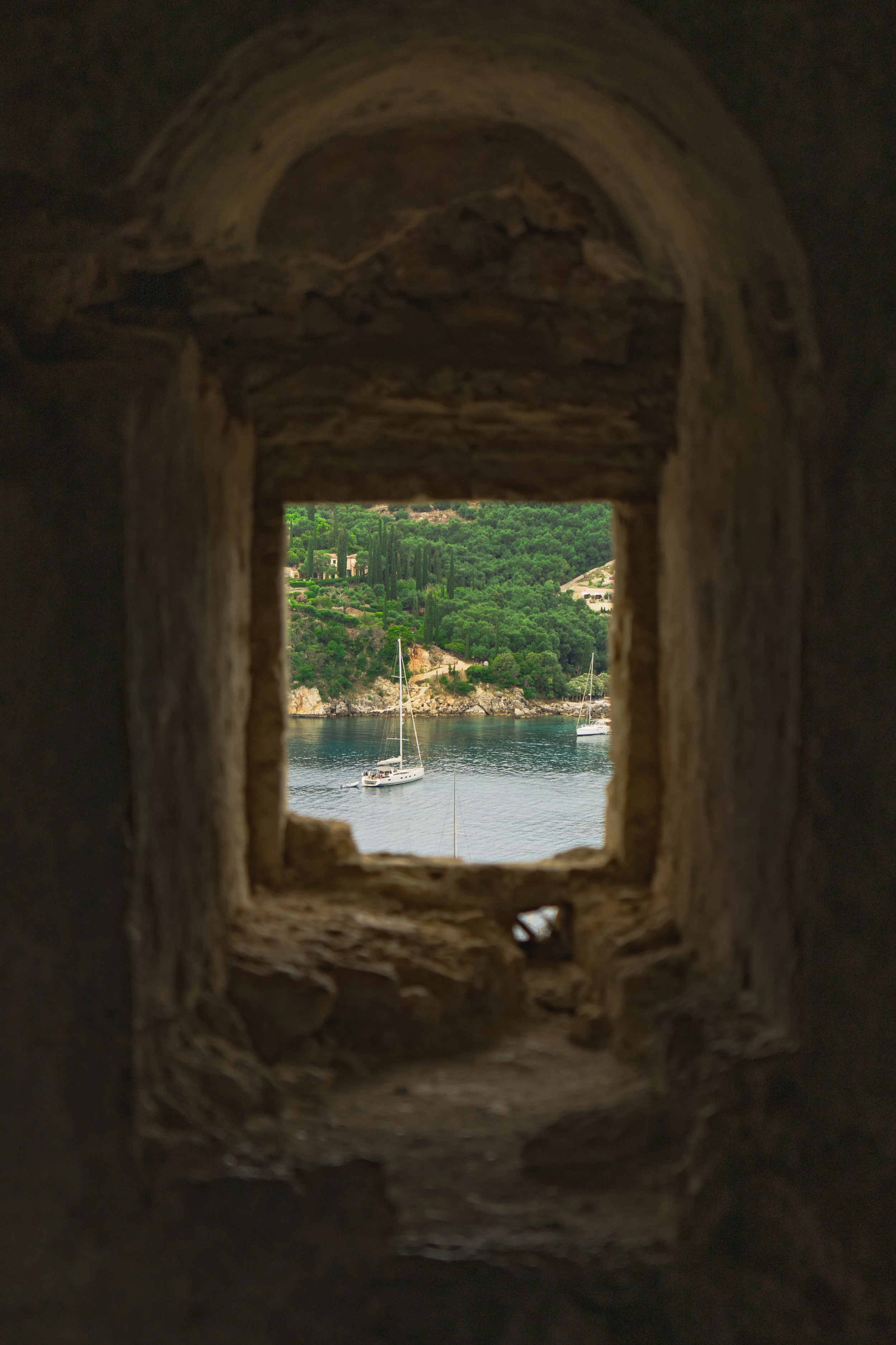 View of a body of water with sailboats and lush green hills in the background, seen through a small rectangular opening in a stone or brick structure.