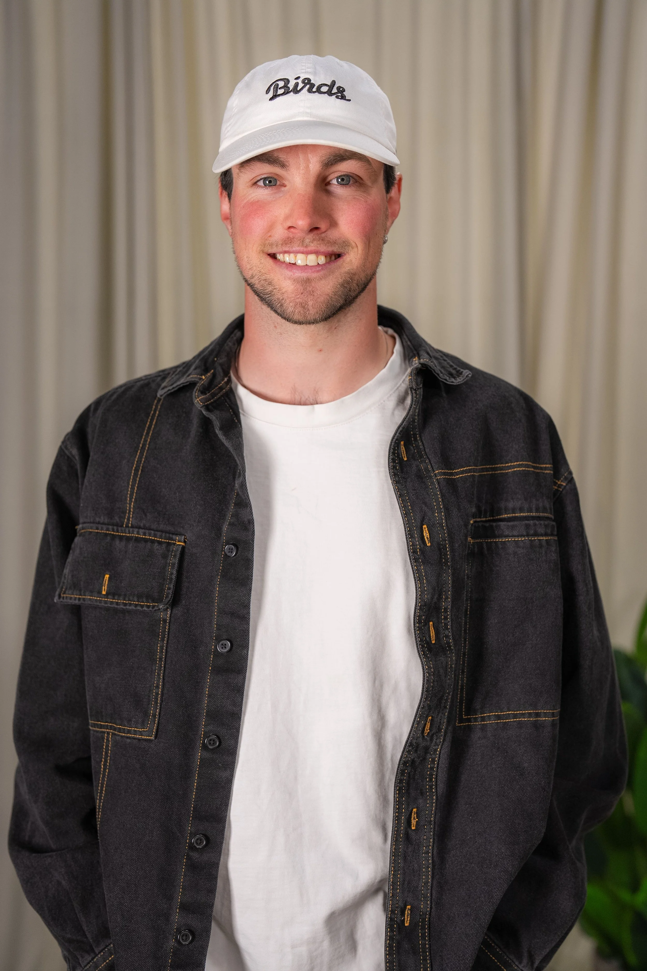 A man with short brown hair and blue eyes smiling, wearing a white cap with 'Birds' embroidered on it, a white t-shirt, and a black denim jacket, standing in front of beige curtains.
