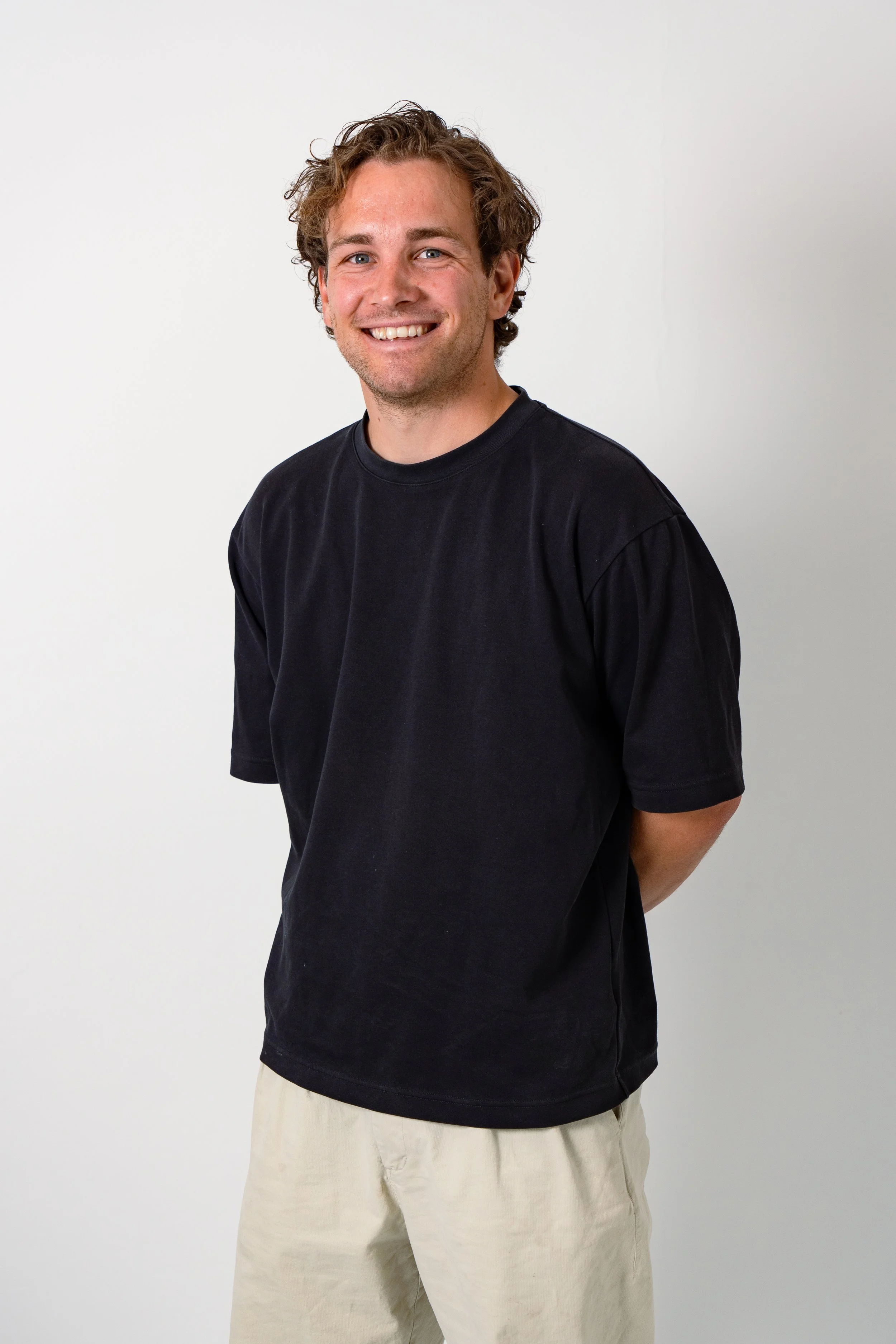 A young man with curly light brown hair, blue eyes, and light facial hair, smiling and wearing a black t-shirt and light-colored pants, standing against a plain white background.