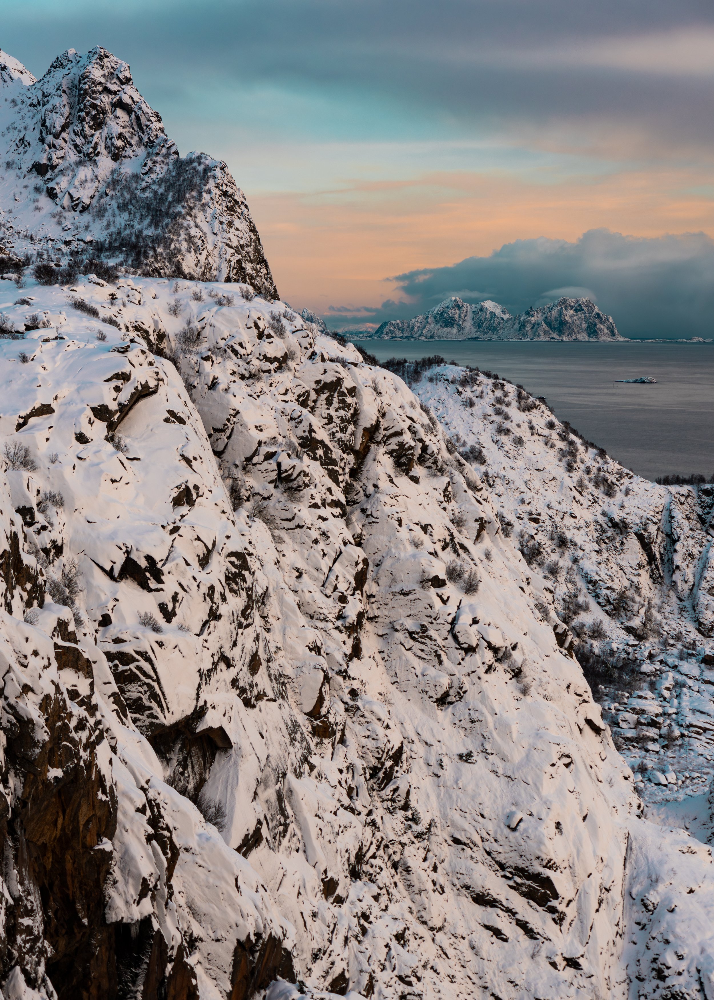 Snow-covered rocky mountain slope with sparse bushes, overlooking a body of water with distant snow-capped mountains and a pastel sky.
