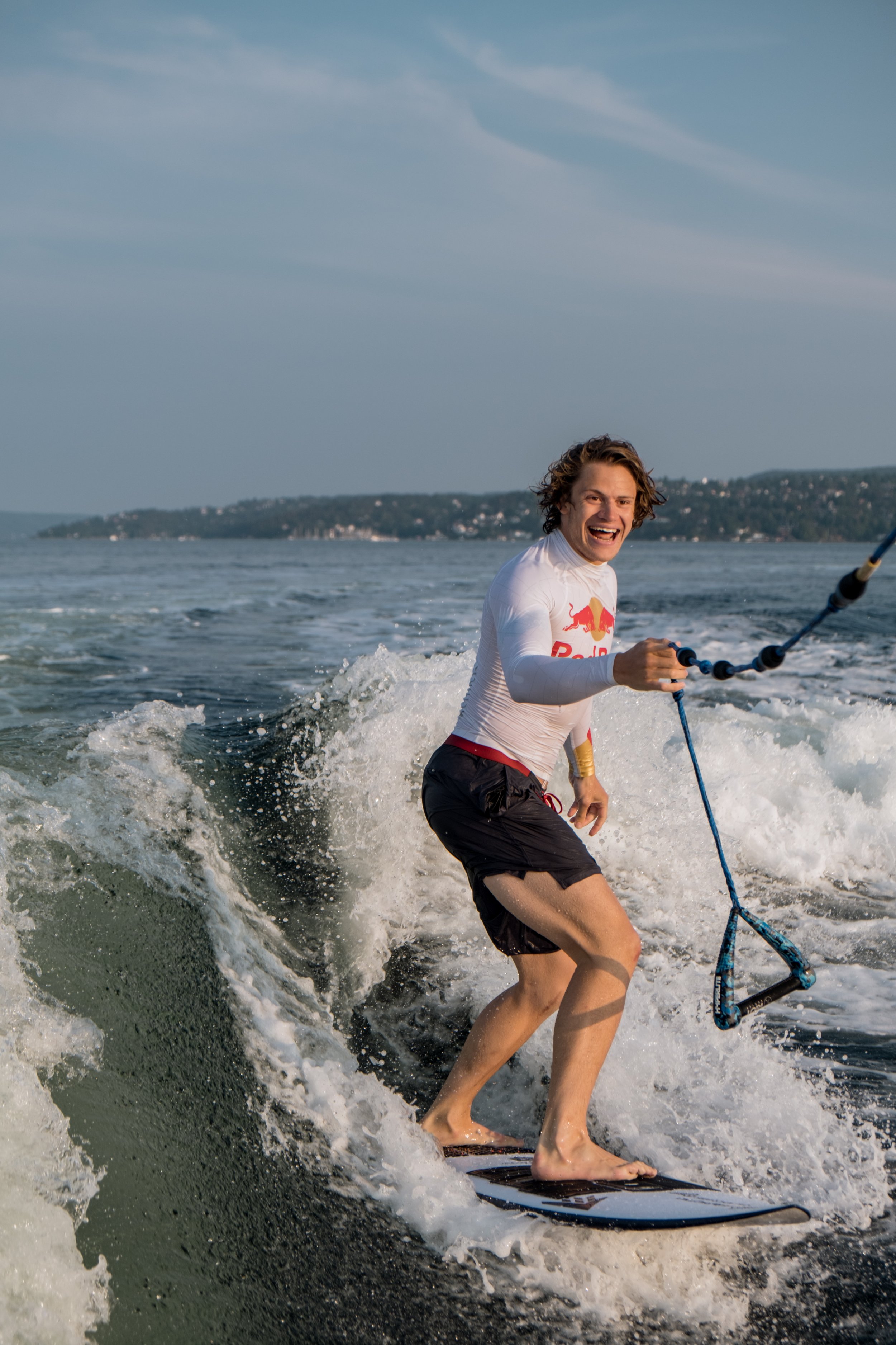 A man wakeboarding on a lake, smiling and holding onto a tow rope, with a distant shoreline and cloudy sky in the background.