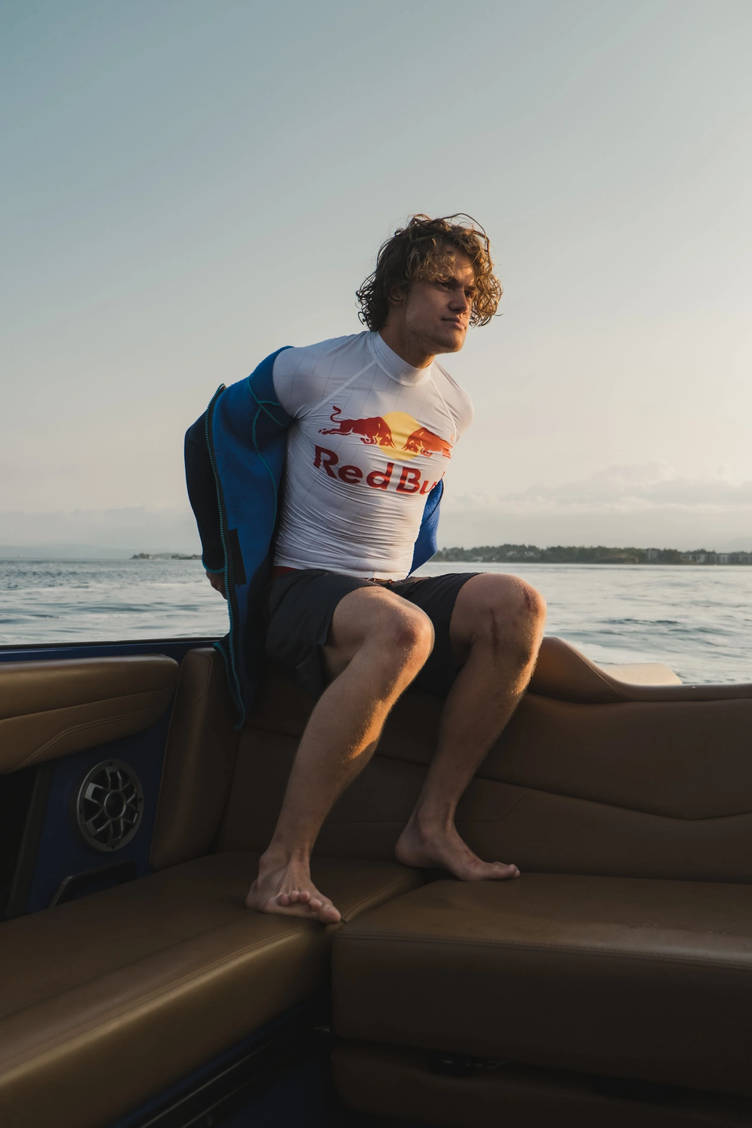 A young man sitting on the couch of a boat, wearing a white Red Bull racing shirt, a blue jacket, and black shorts. He is barefoot, with his hands behind his back and leaning forward slightly. The water and a distant shoreline are visible in the back