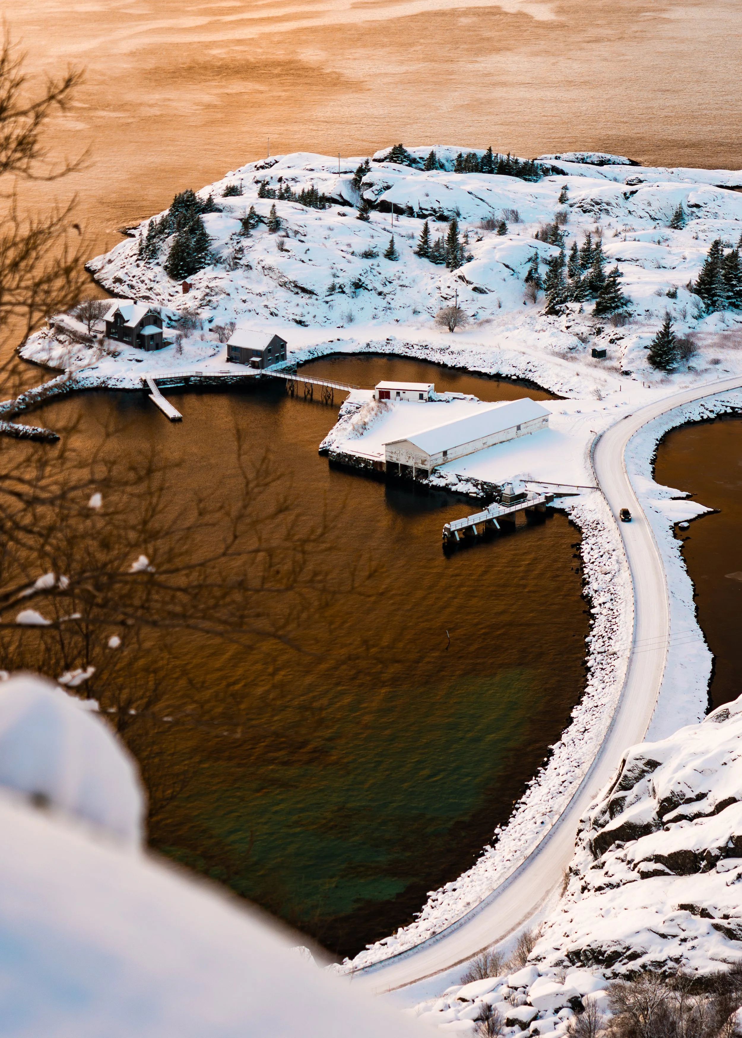 Snow-covered rocky island with trees, a small bridge, houses, and a curved road, viewed through an airplane window at sunset.