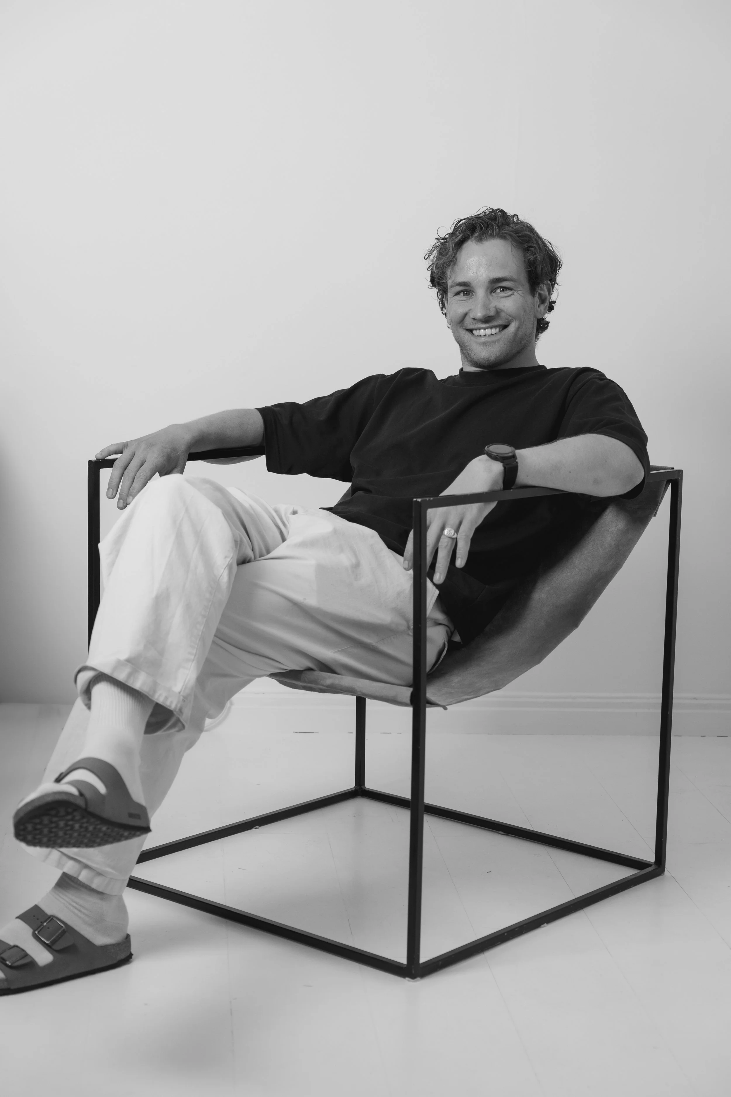 A smiling man with curly hair sitting comfortably in a modern chair with a metal frame, wearing casual clothing and sneakers, in a minimalistic indoor setting.