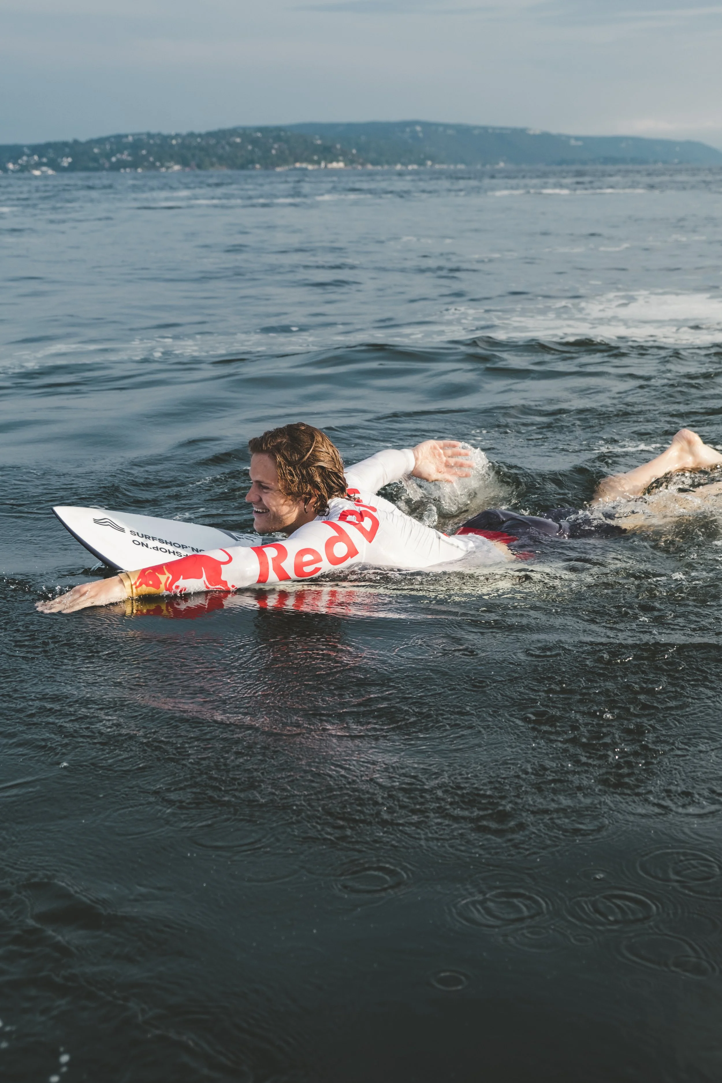 A surfer with long hair and a broad smile paddles on a surfboard wearing a white rash guard with red logo and text on the arm, in the ocean with a distant landmass or island.