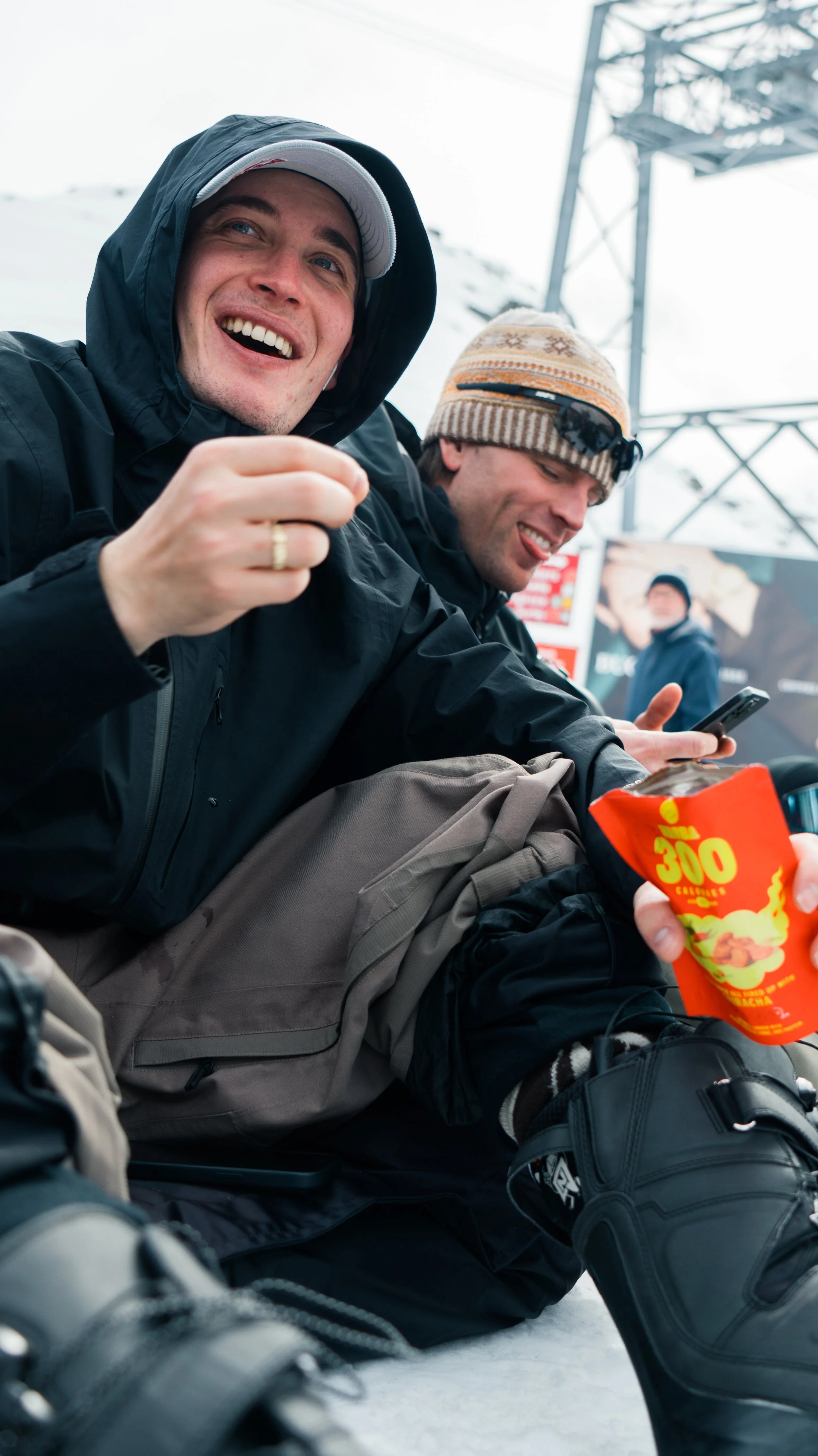 Two men sitting outdoors in snowy weather, wearing winter clothing, smiling, and enjoying snacks. One man is wearing a black jacket and hood, and the other is wearing a brown and beige knit cap with sunglasses.
