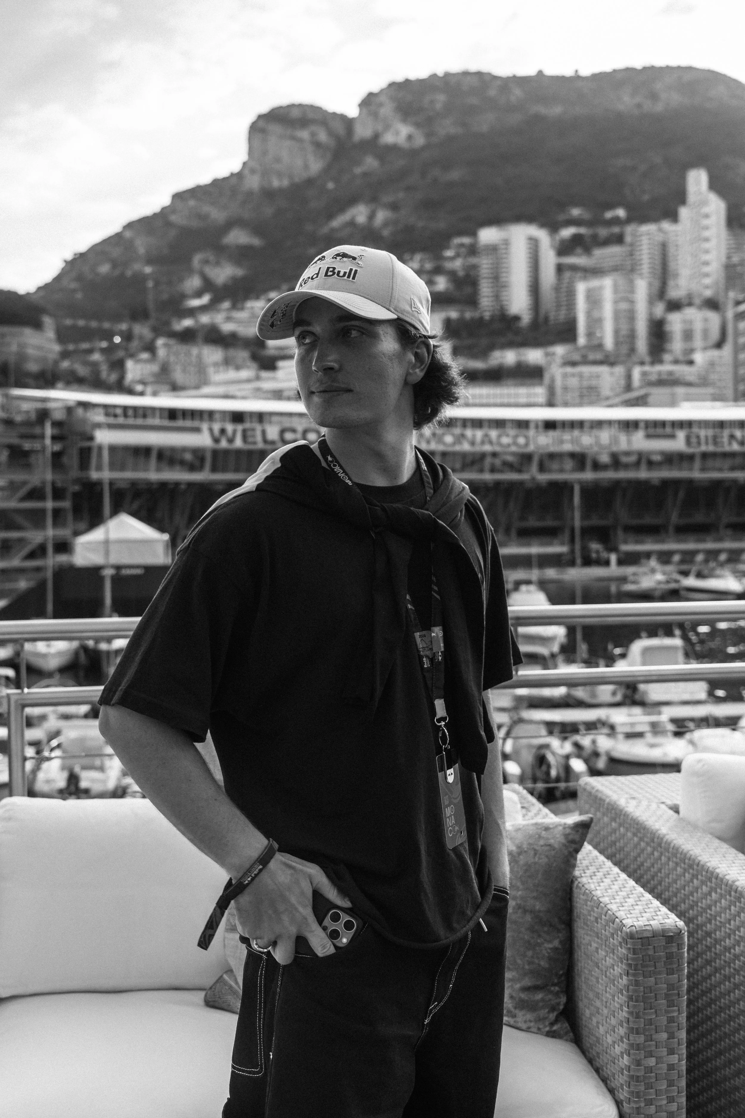 A young man wearing a baseball cap with the Red Bull logo, standing outdoors with a mountain and cityscape in the background, holding a smartphone in his right hand.
