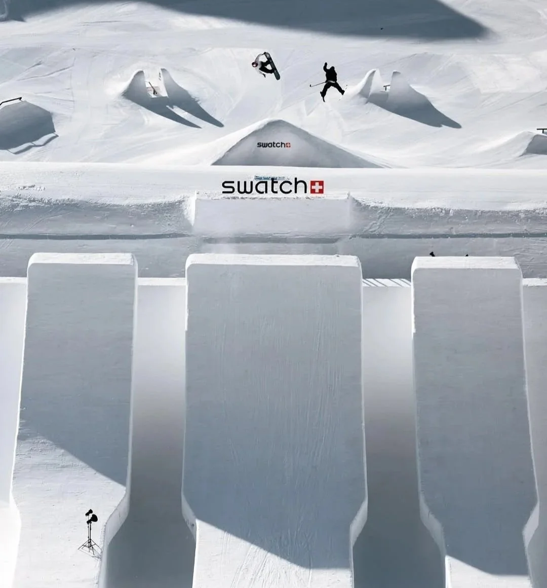 Snowboarder and skier performing tricks on a snow terrain park with ramps and jumps, with a Swatch logo on the snow.
