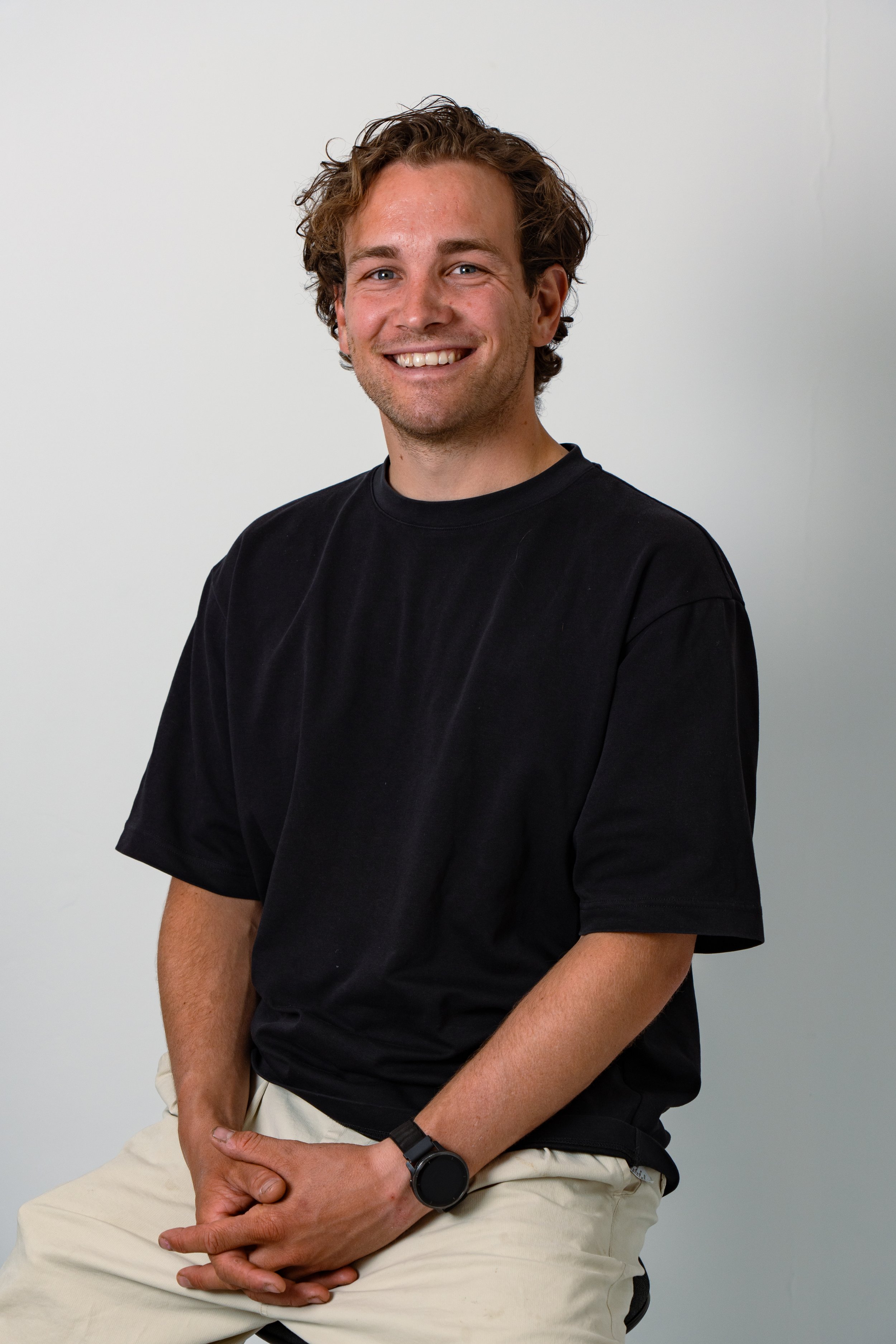 A young man with curly brown hair, wearing a black t-shirt and beige pants, sitting against a plain white wall, smiling at the camera.