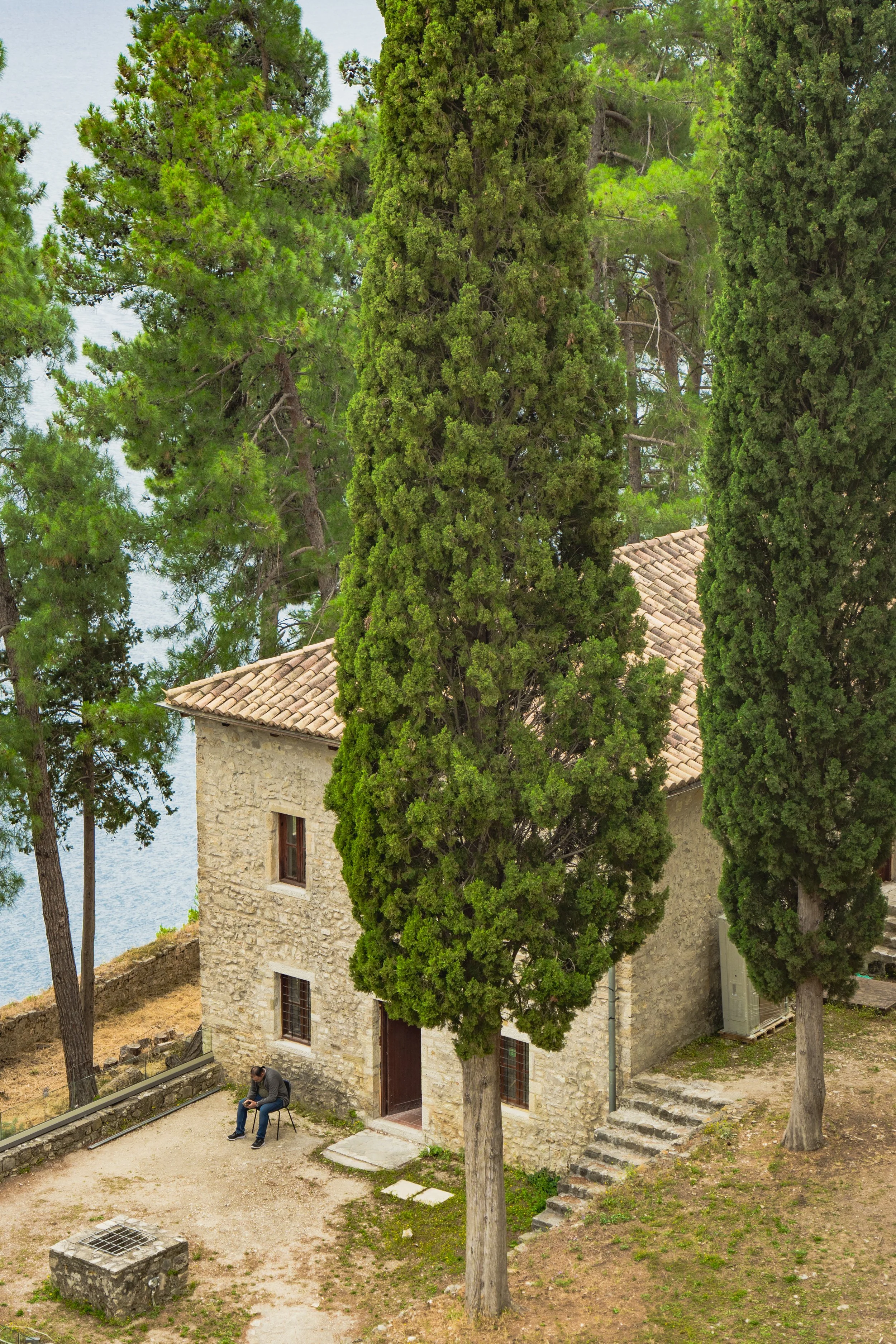 A stone building with a tiled roof, surrounded by tall green trees, with a man sitting on a bench outside, near a river.