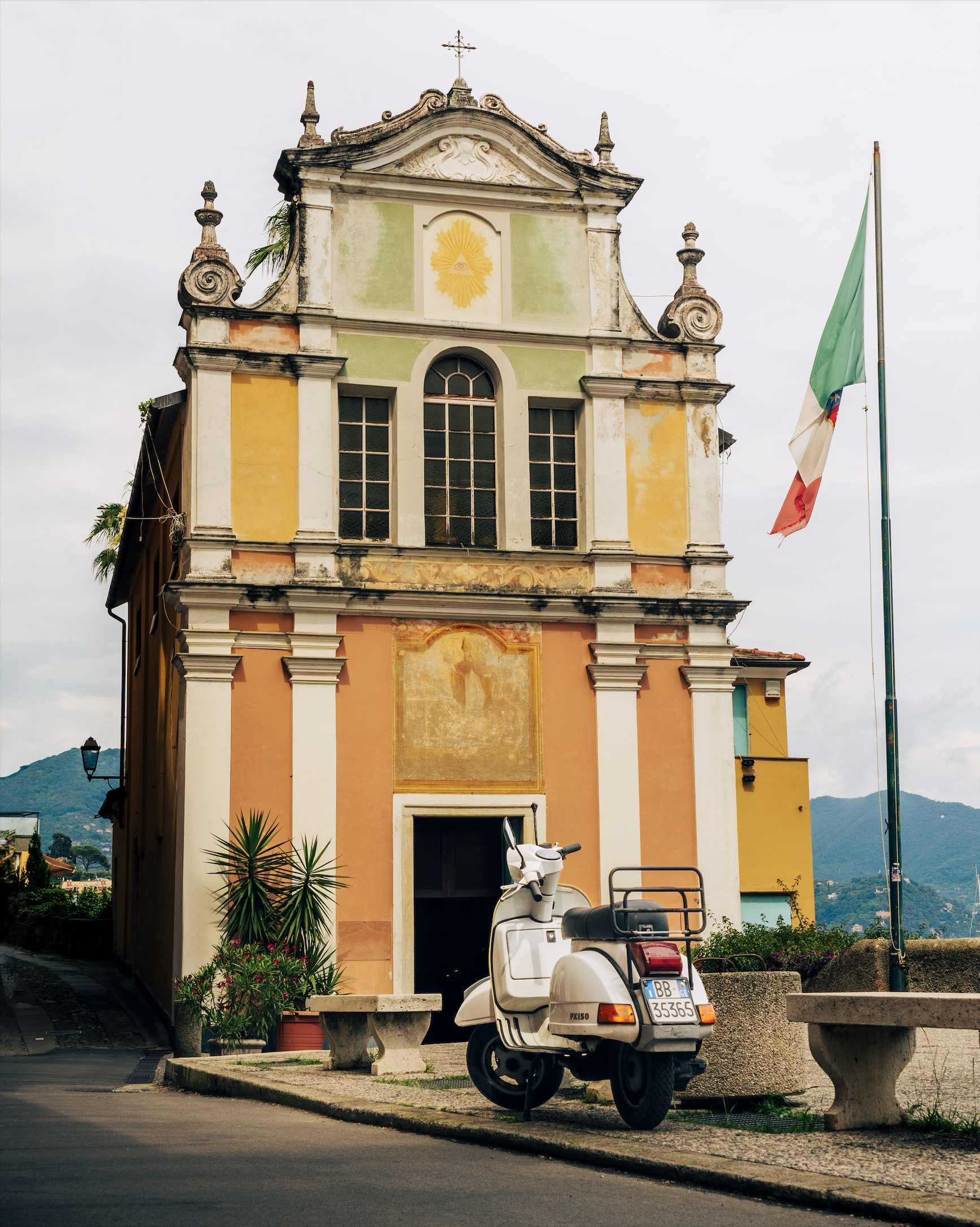 Colorful historical building with a small scooter parked in front, surrounded by plants and a flagpole with an Italian flag against a cloudy sky.