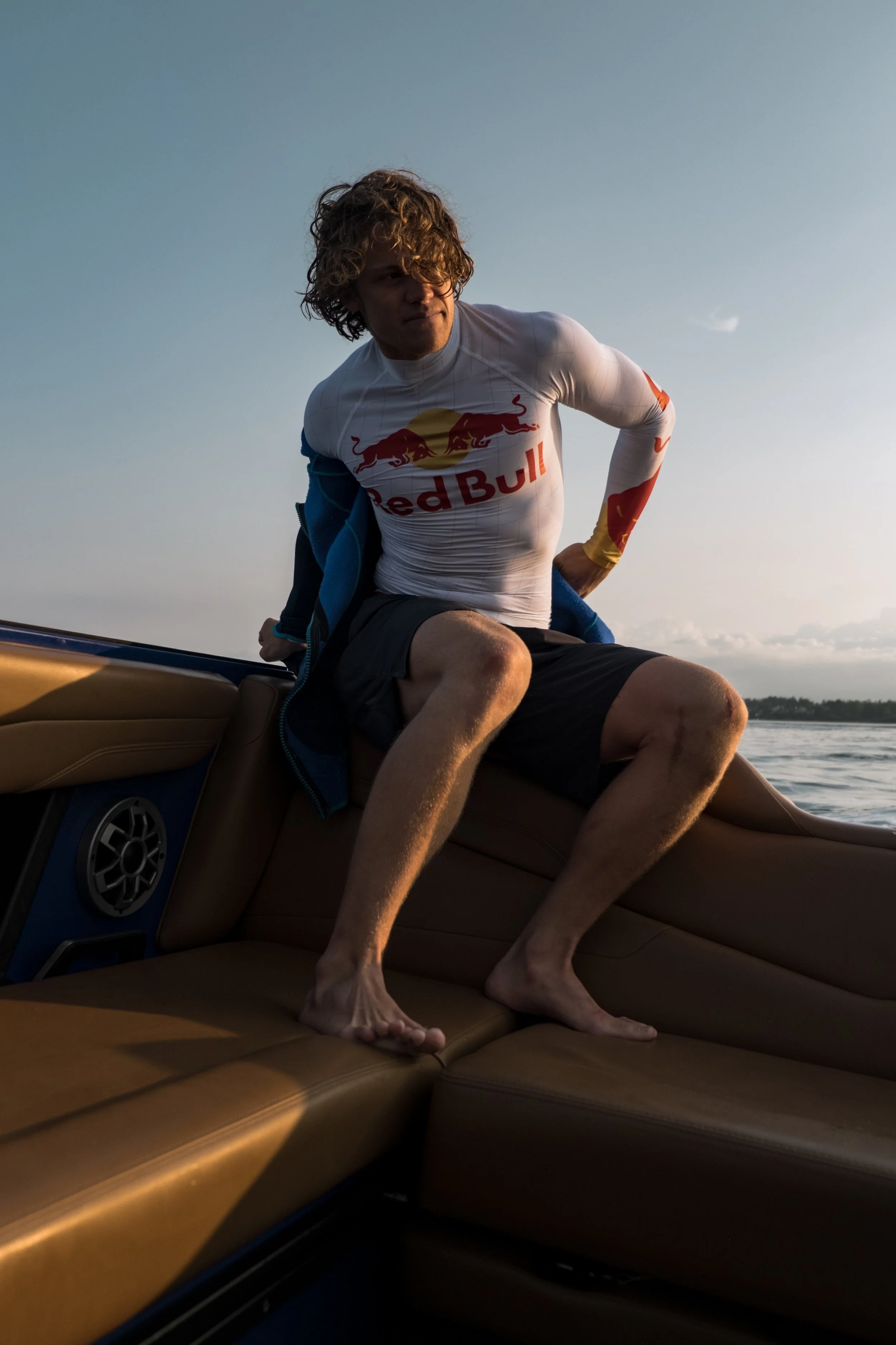 A man with curly hair and wearing a Red Bull racing shirt is sitting on the leather seats of a boat, with his legs stretched out and feet resting on the seat. The boat is on a body of water, with a partly cloudy sky in the background.