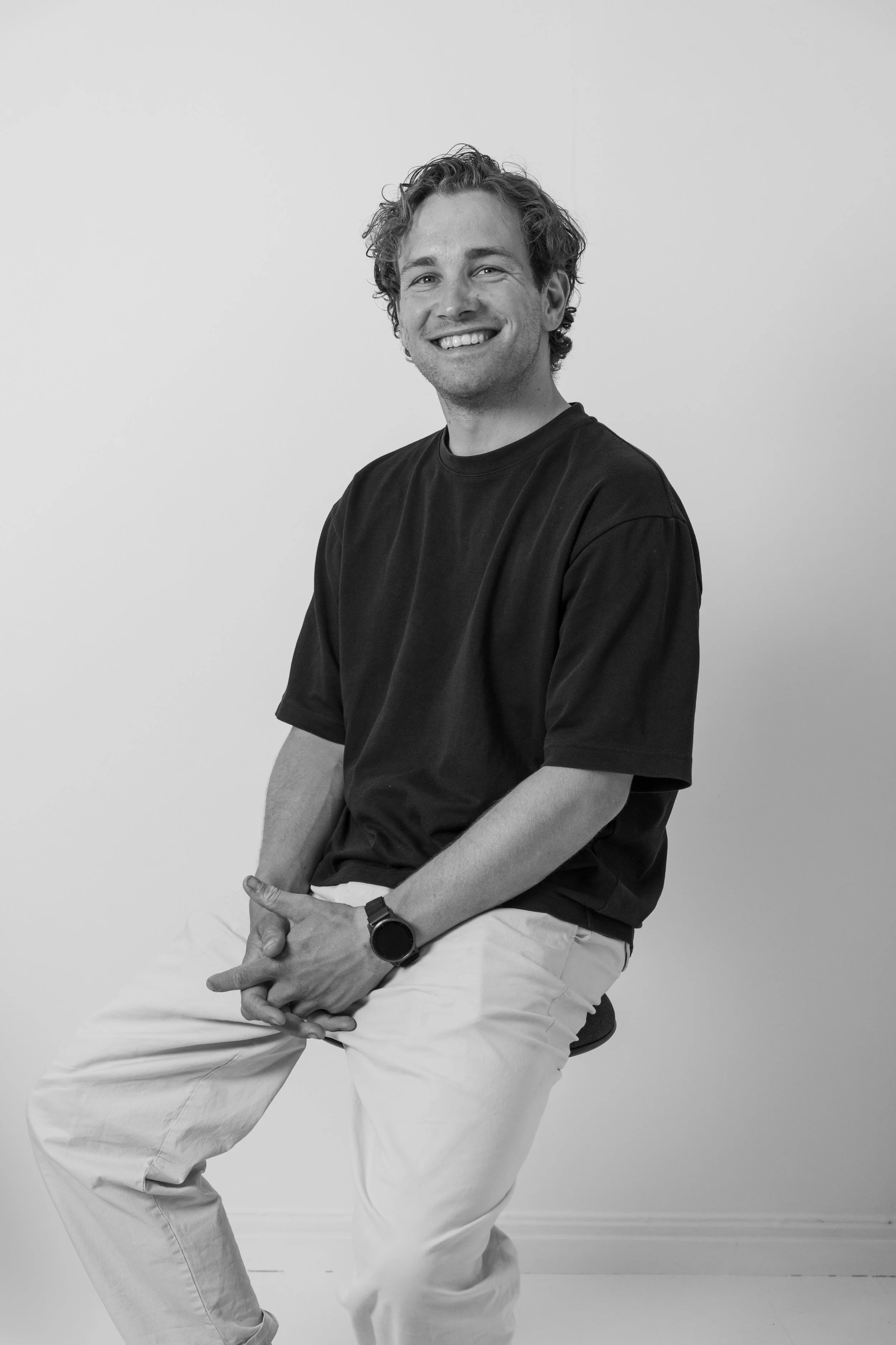 A young man with curly hair smiling and sitting on a stool against a plain wall, wearing a black t-shirt, white pants, and a watch.