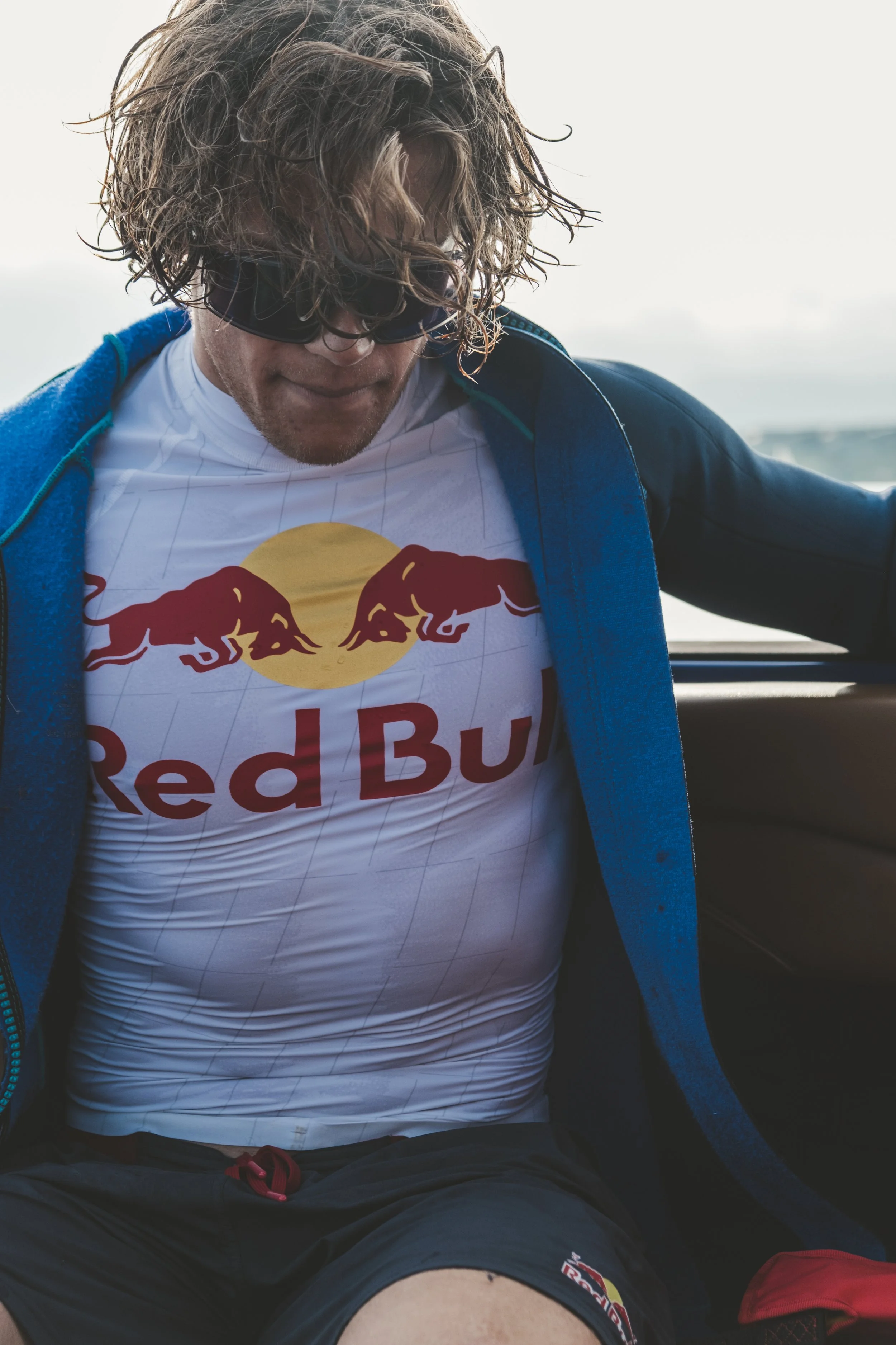 A man with curly hair, wearing sunglasses, a white and red Red Bull t-shirt, black shorts, and a blue hoodie, sitting outdoors during daytime.
