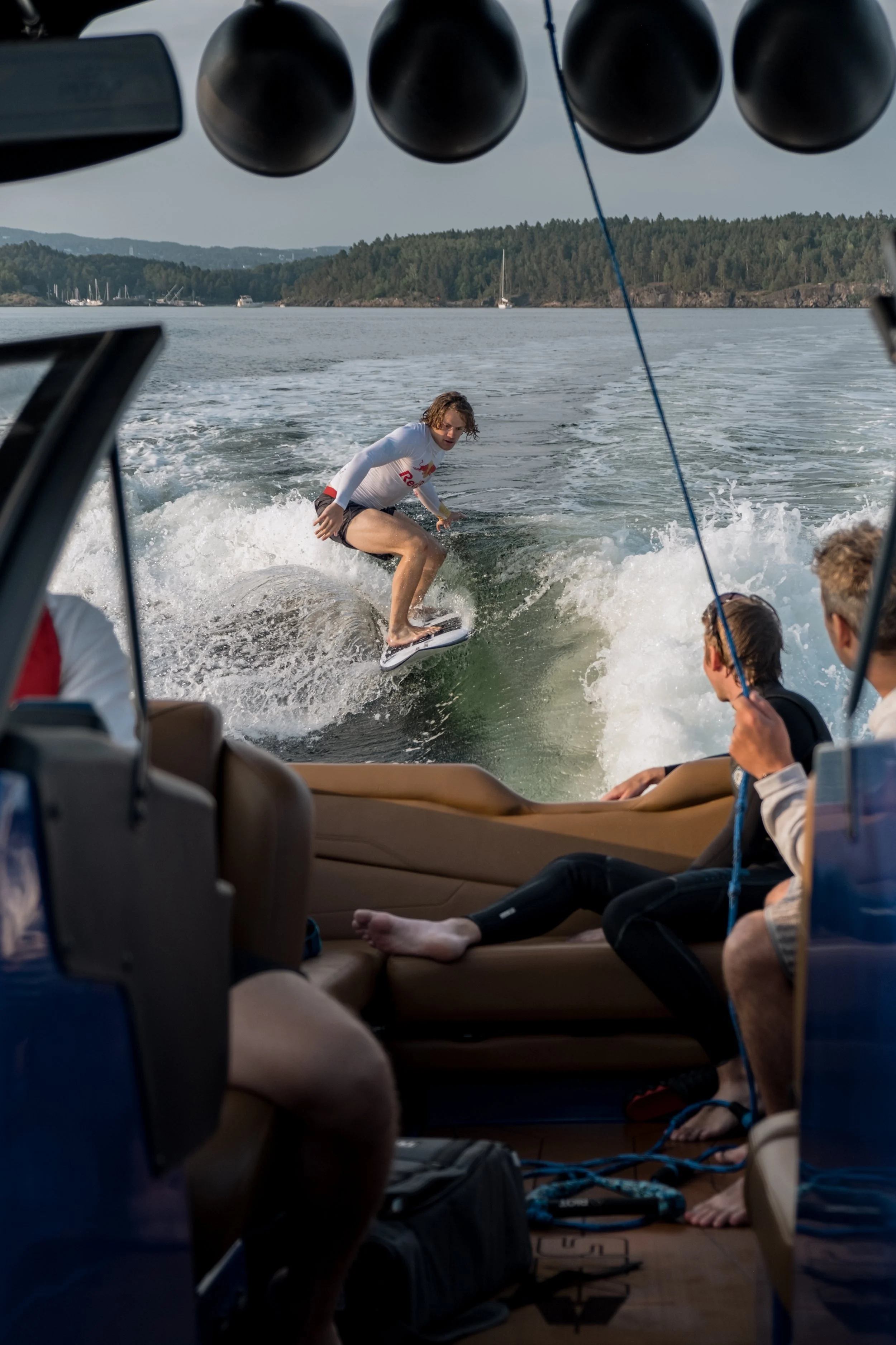 A person surfing on a wakeboard behind a boat on a body of water, seen from the boat's interior with others sitting onboard.