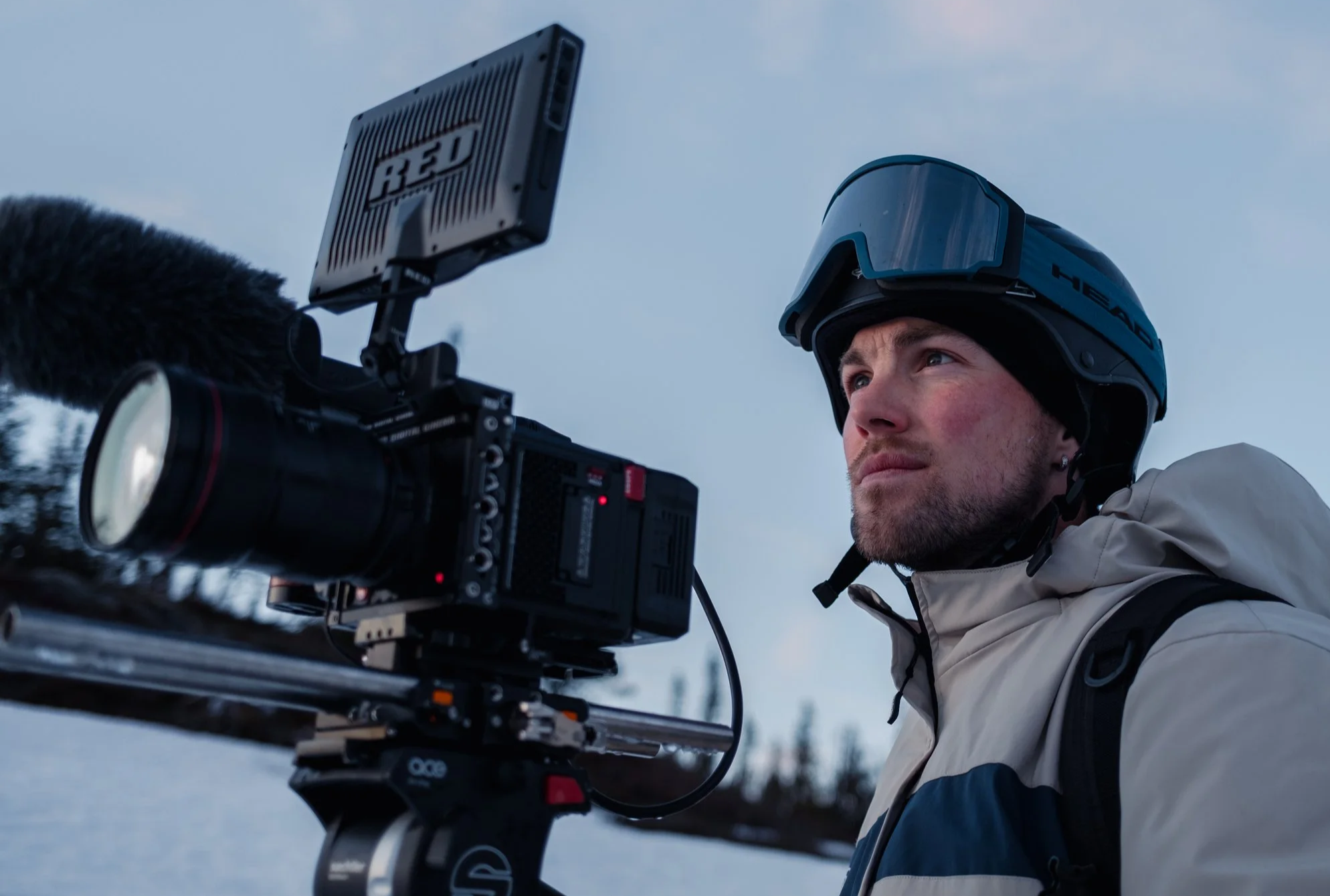 A man wearing a helmet and winter gear operating a professional camera mounted on a tripod outdoors in a snowy landscape.