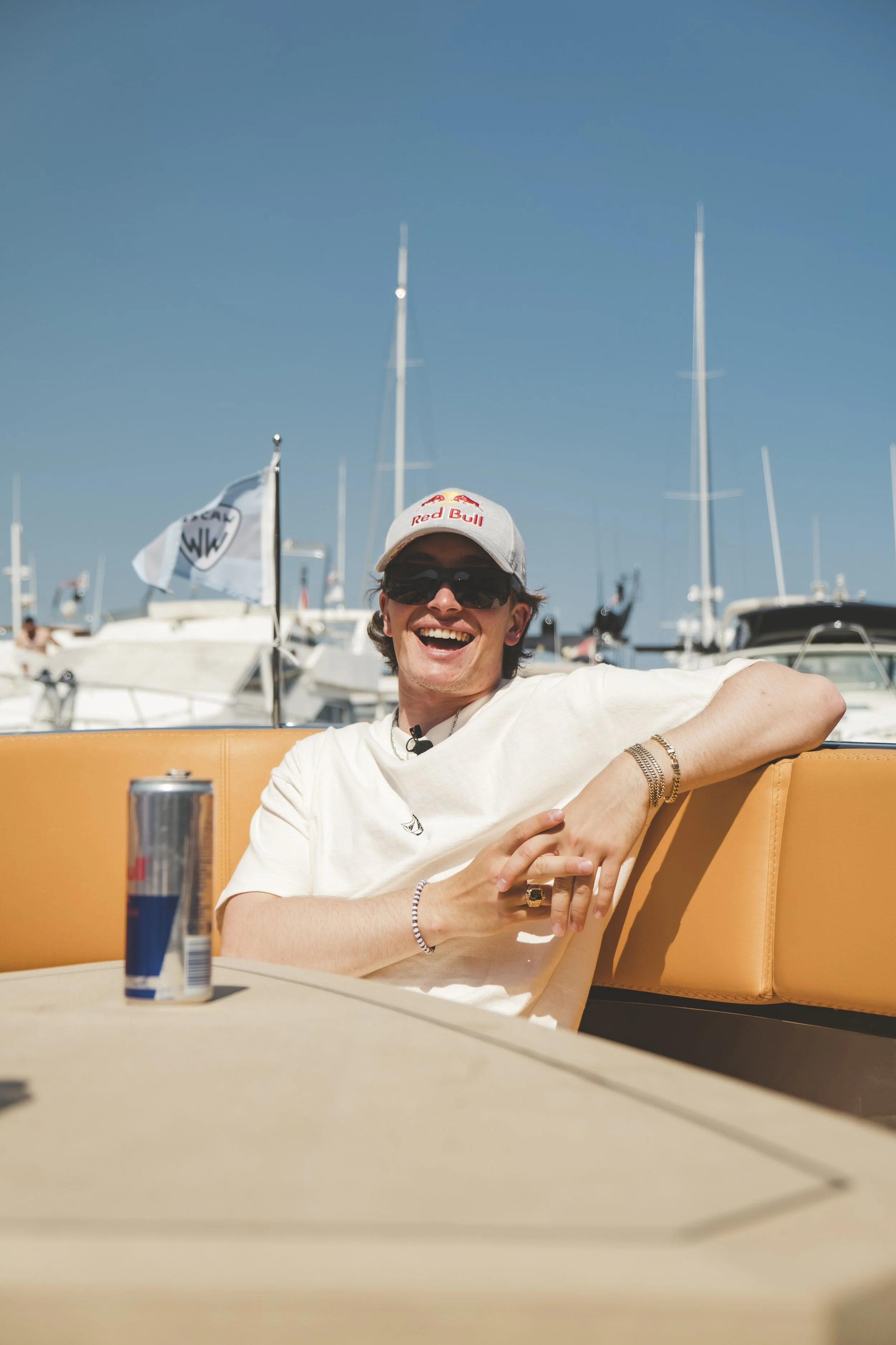 A young man wearing sunglasses, a gray Red Bull cap, and a white t-shirt is sitting on a yellow bench outdoors at a marina with boats and masts in the background. He is smiling and gesturing with his left hand, with a can of Red Bull on the table in 