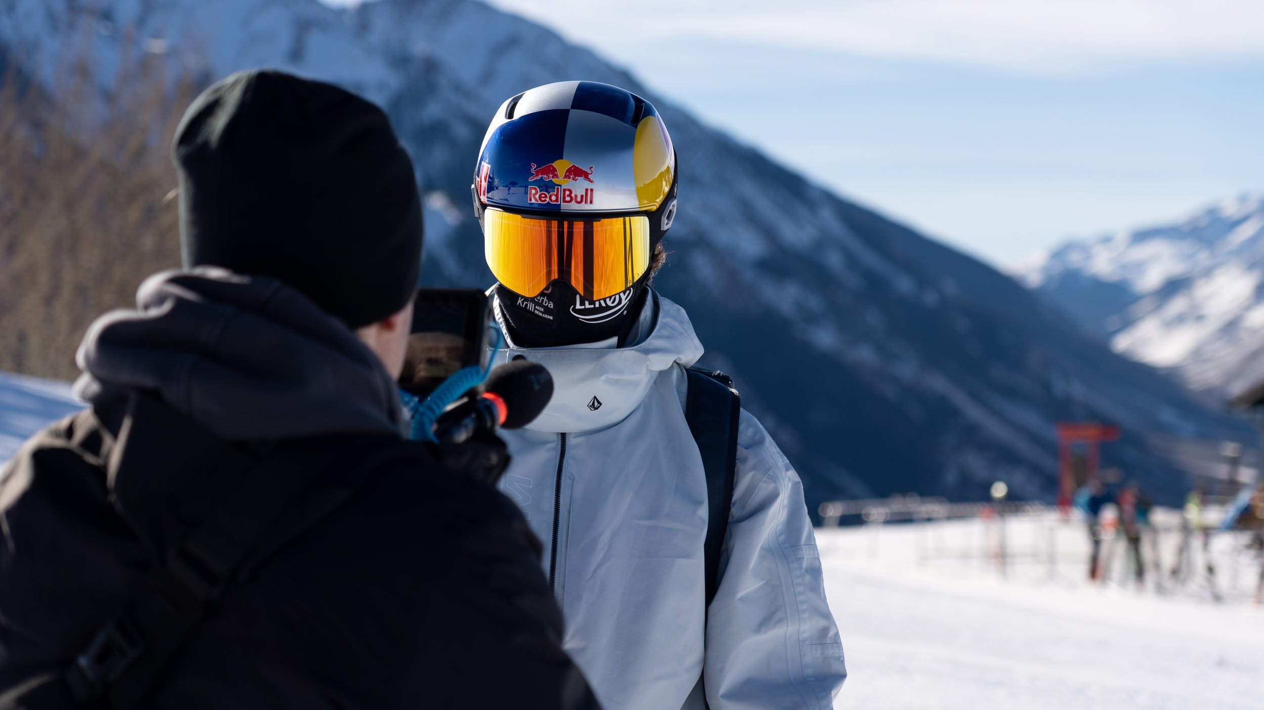 A skier wearing a helmet with Red Bull logo and reflective goggles being interviewed by a person in winter clothing in a snowy mountain landscape.