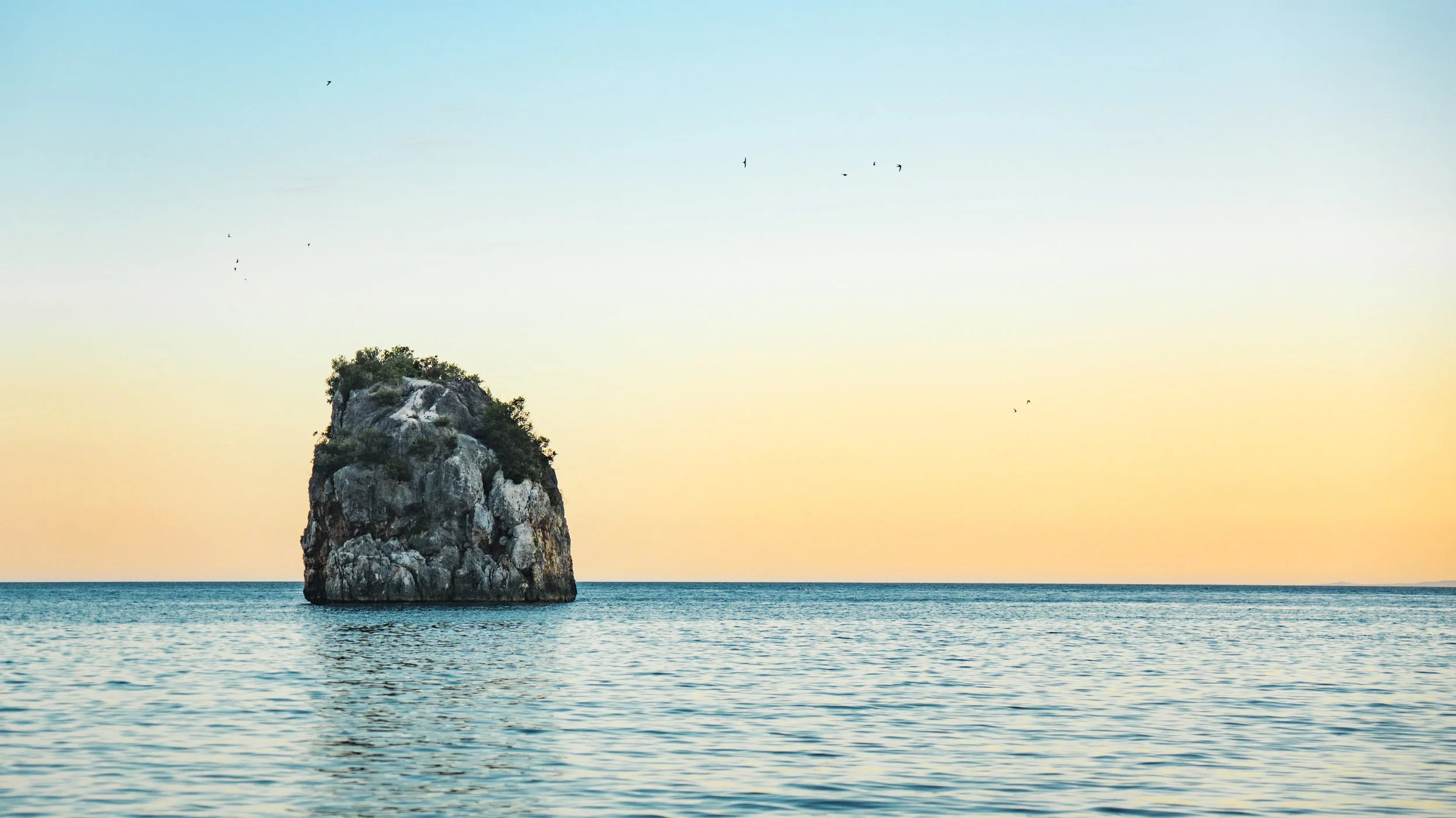 A large rock formation with greenery on top is in the ocean, with a calm sea and a pastel-colored sky during sunset or sunrise, and birds flying overhead.