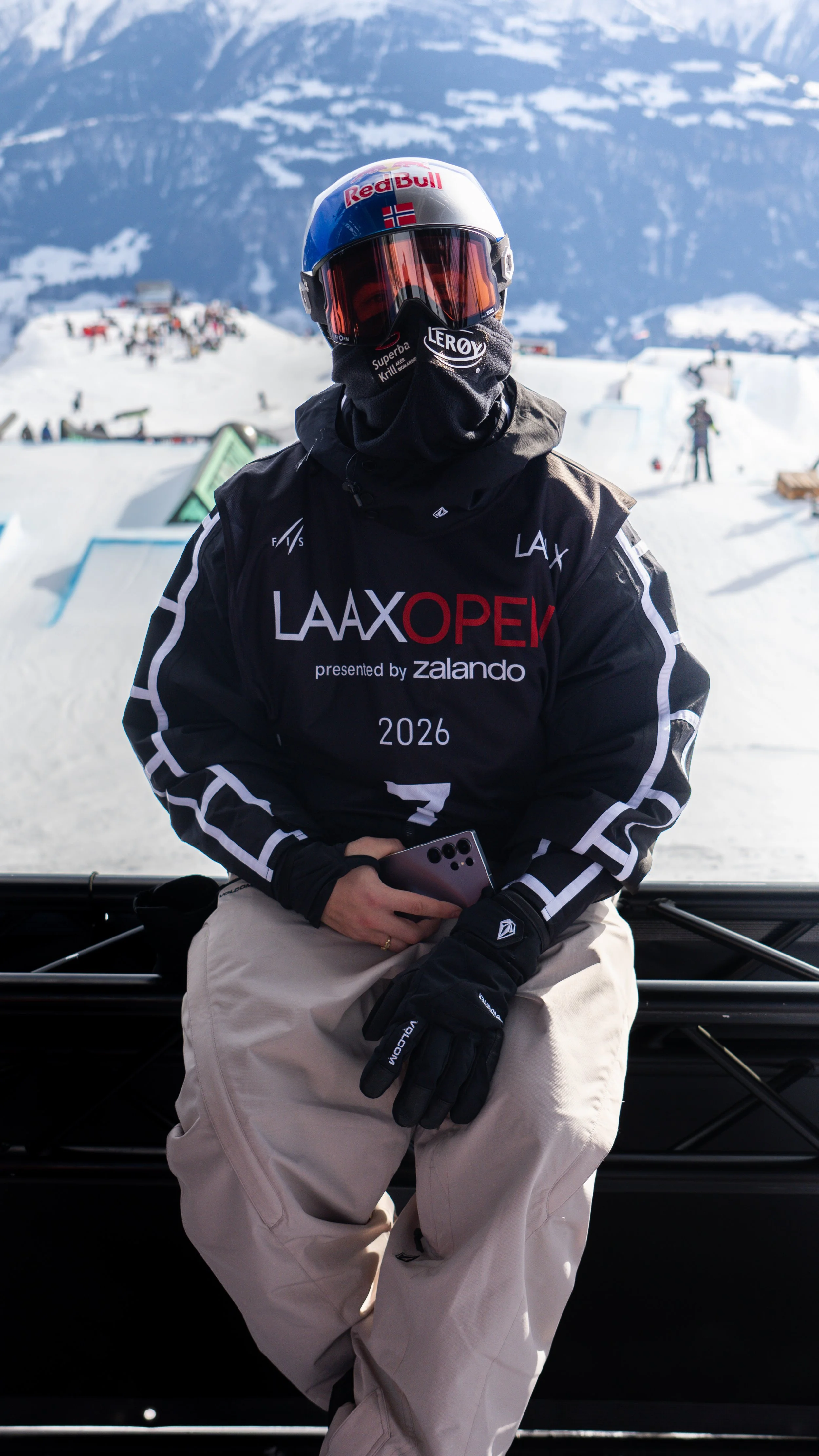 A person wearing winter sports gear, including a Red Bull helmet and goggles, sits on a bench in a snowy mountain area with snowboarding and skiing activity in the background.