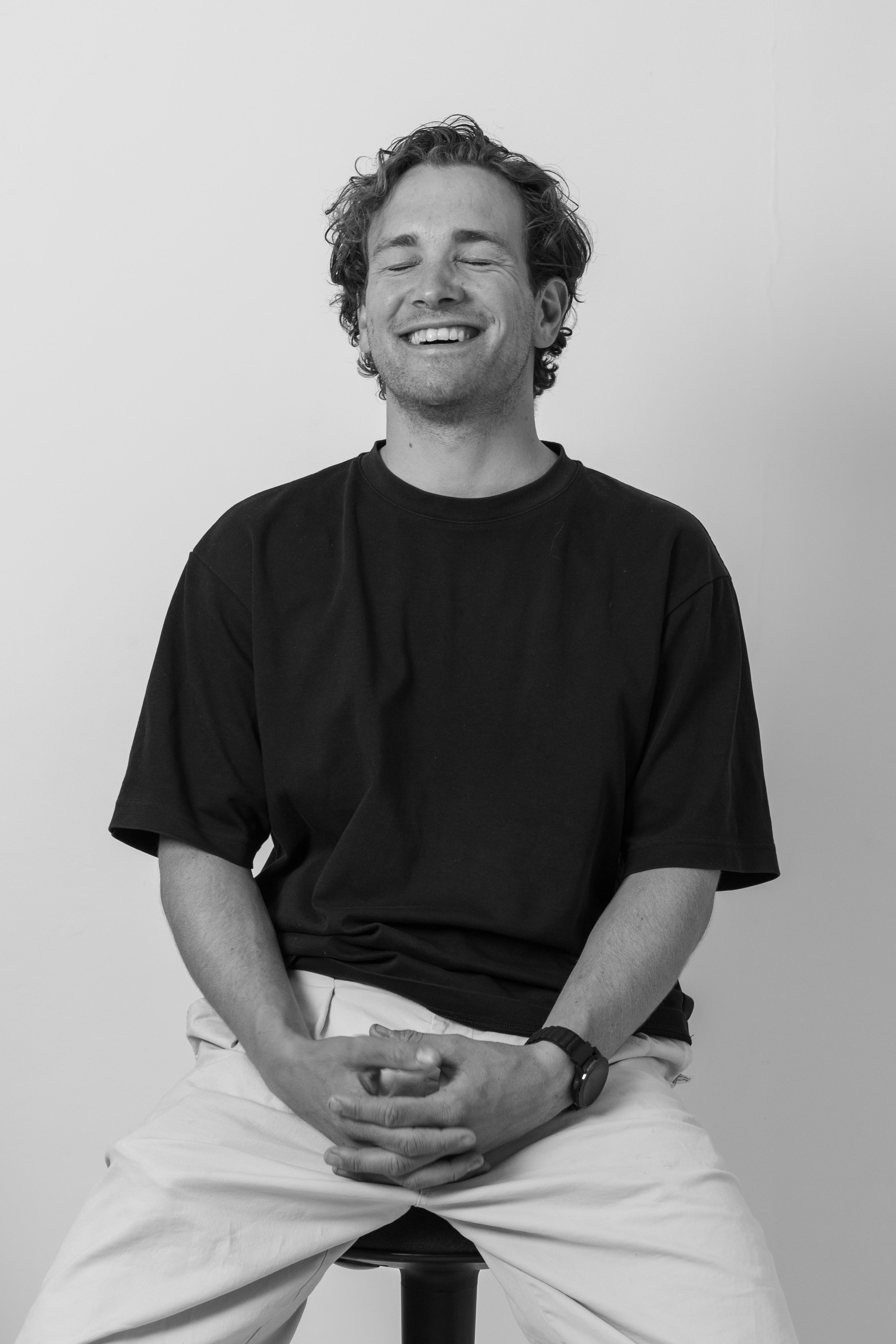 Black and white photo of a man smiling with eyes closed, sitting on a stool, wearing a black t-shirt and white pants, with his hands in his lap and a watch on his left wrist.
