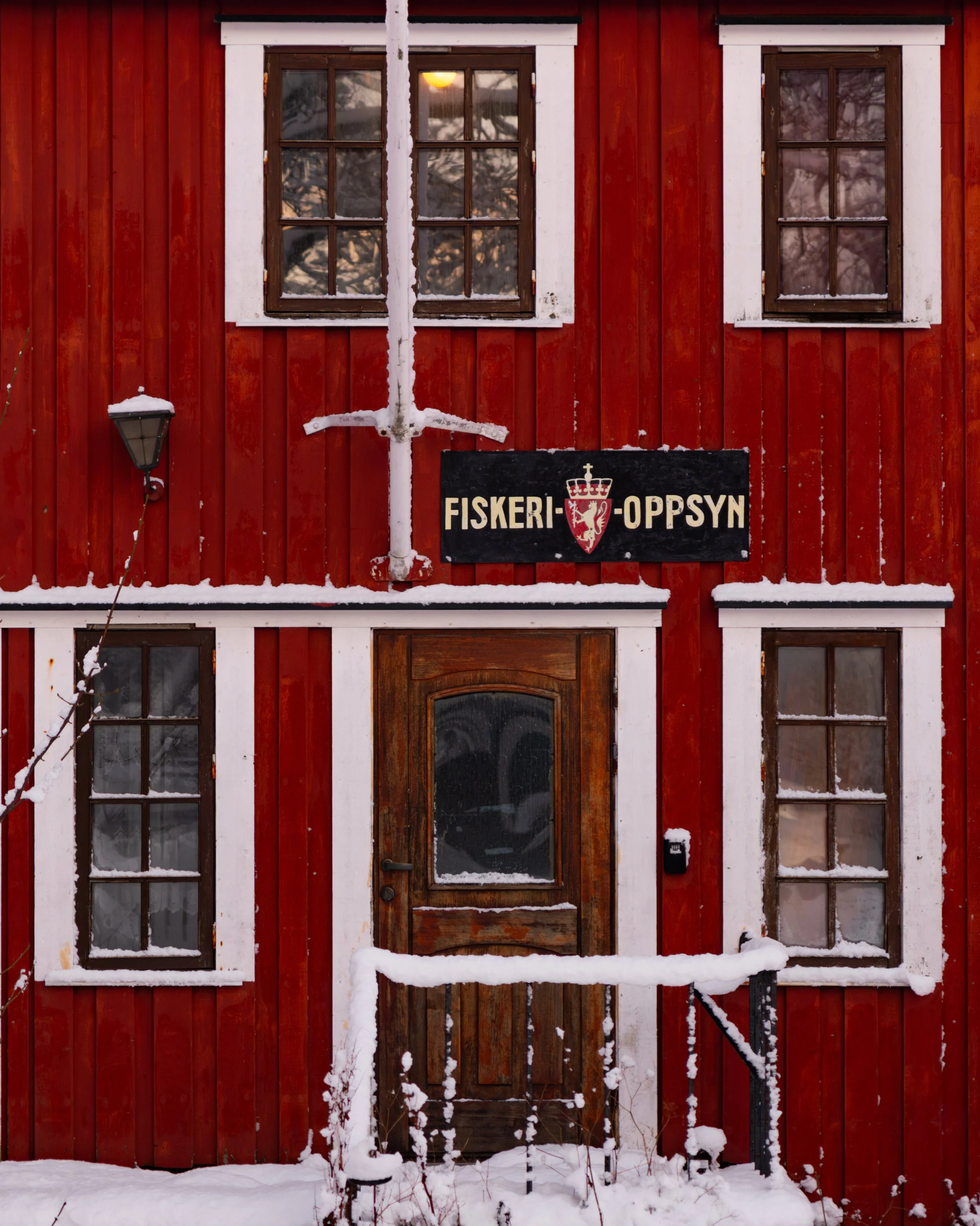 Red wooden building with two upper windows, a wooden door in the center, snow on the ground, and a sign reading 'Fiskeri Oppsyn' with a shield emblem. Snow covers some structures and the sign's top edge.