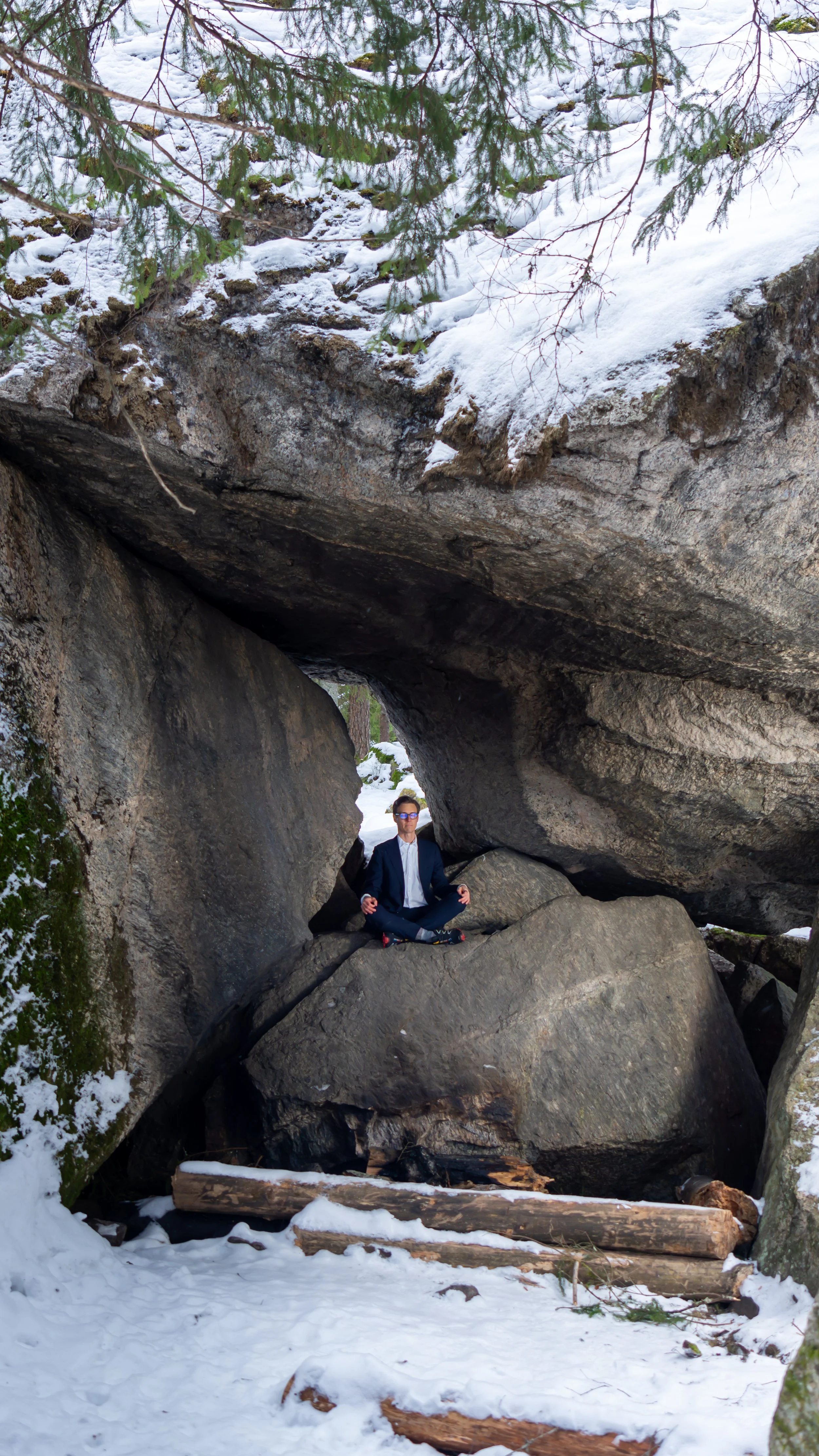A person in a suit sitting cross-legged and meditating inside a natural rock formation, surrounded by snow and trees.
